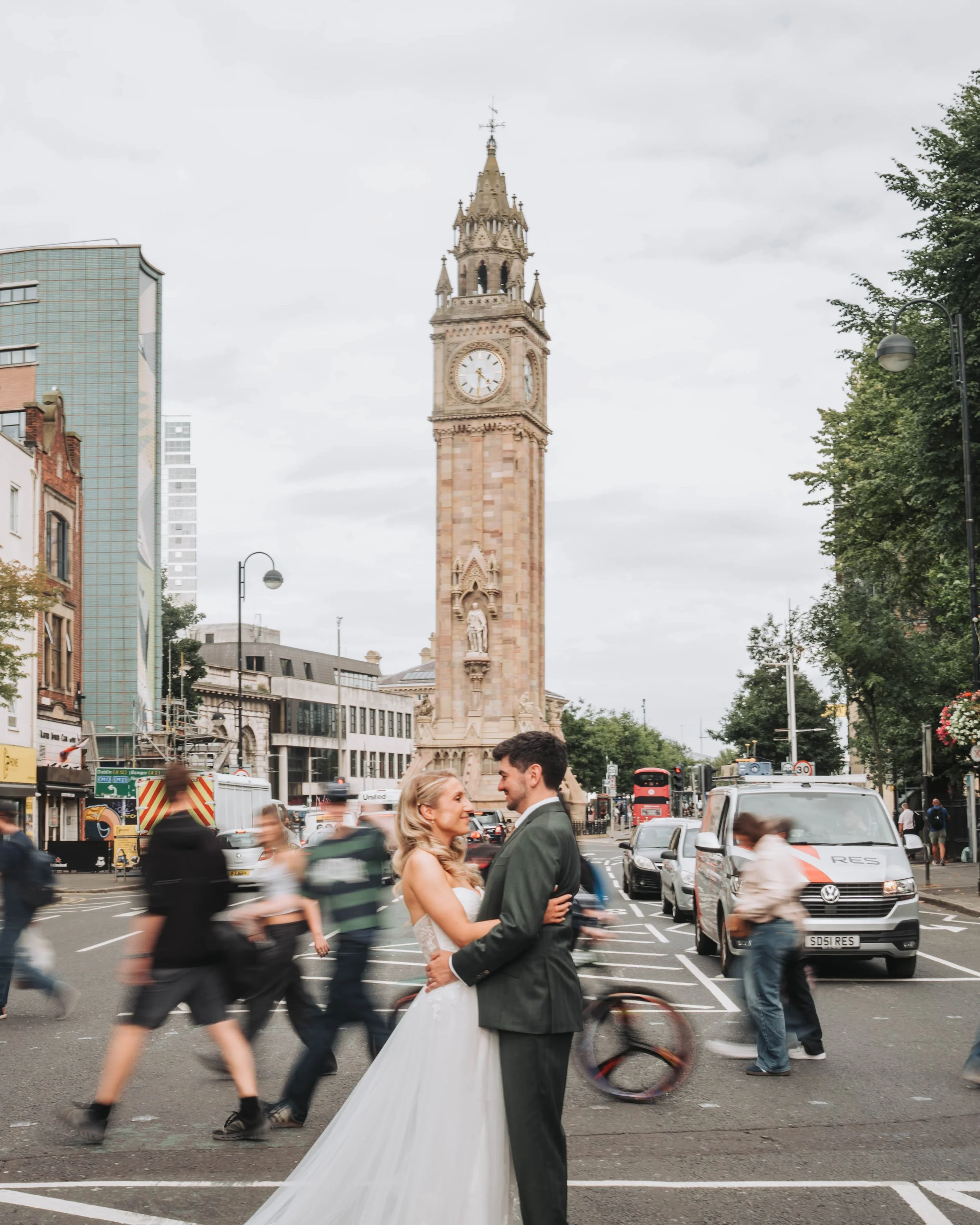 A newlywed couple in wedding attire dancing or embracing on a busy city street with blurred pedestrians and vehicles, and a historic clock tower in the background.