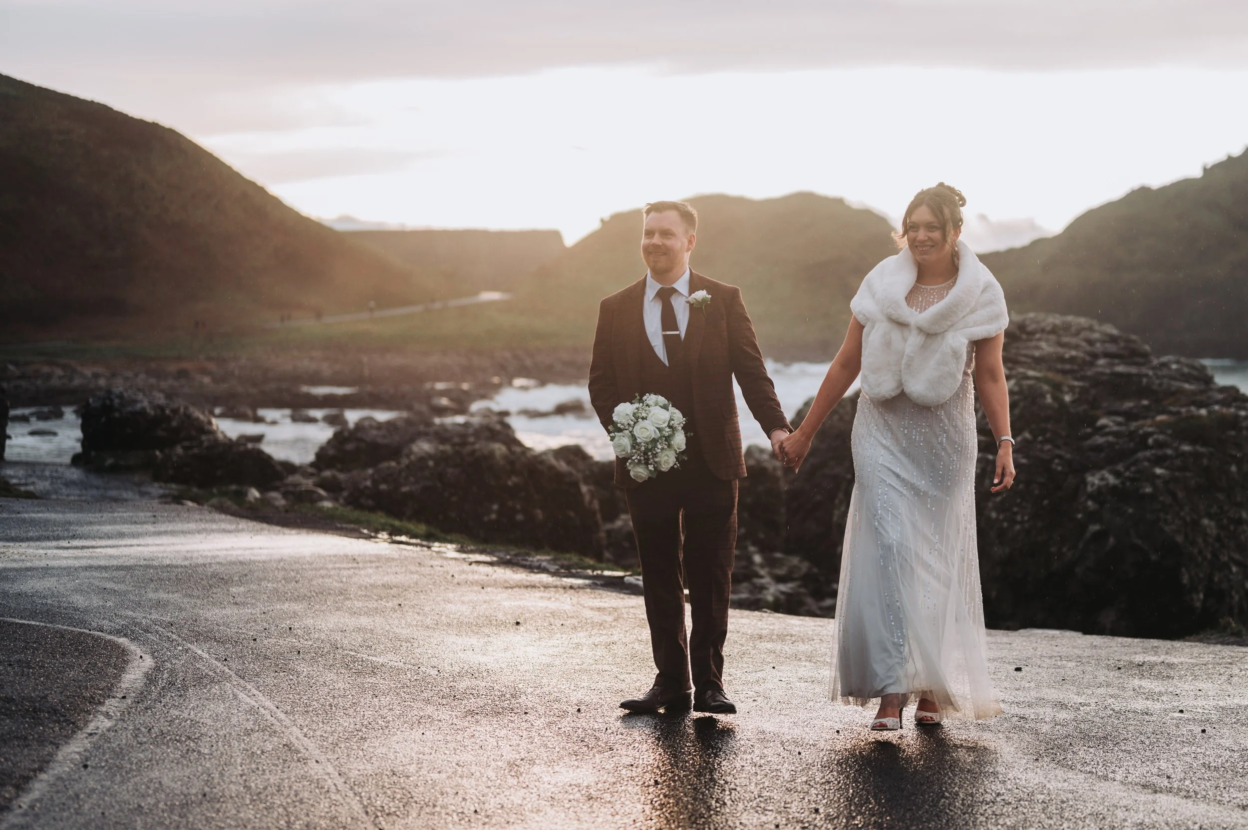 A newlywed couple walking hand-in-hand on a wet road beside the ocean at sunset. The groom is holding a bouquet and wearing a brown suit, the bride is wearing a white dress with a fur stole.