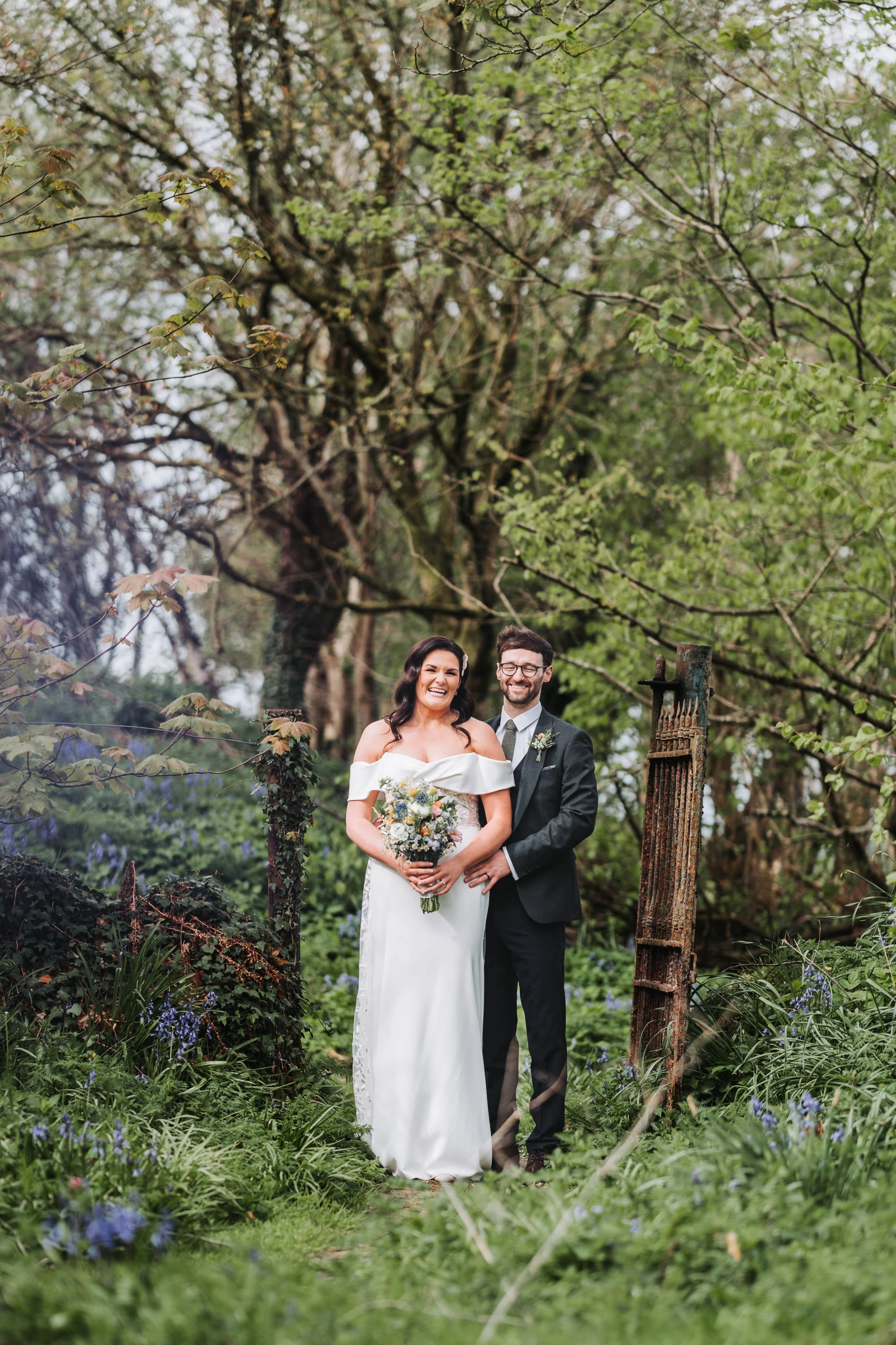 A bride and groom standing together outdoors in a lush garden, smiling and posing for their wedding photo. The bride wears a white off-the-shoulder dress and holds a bouquet, while the groom wears a dark suit with a boutonnière. Tall trees and greene