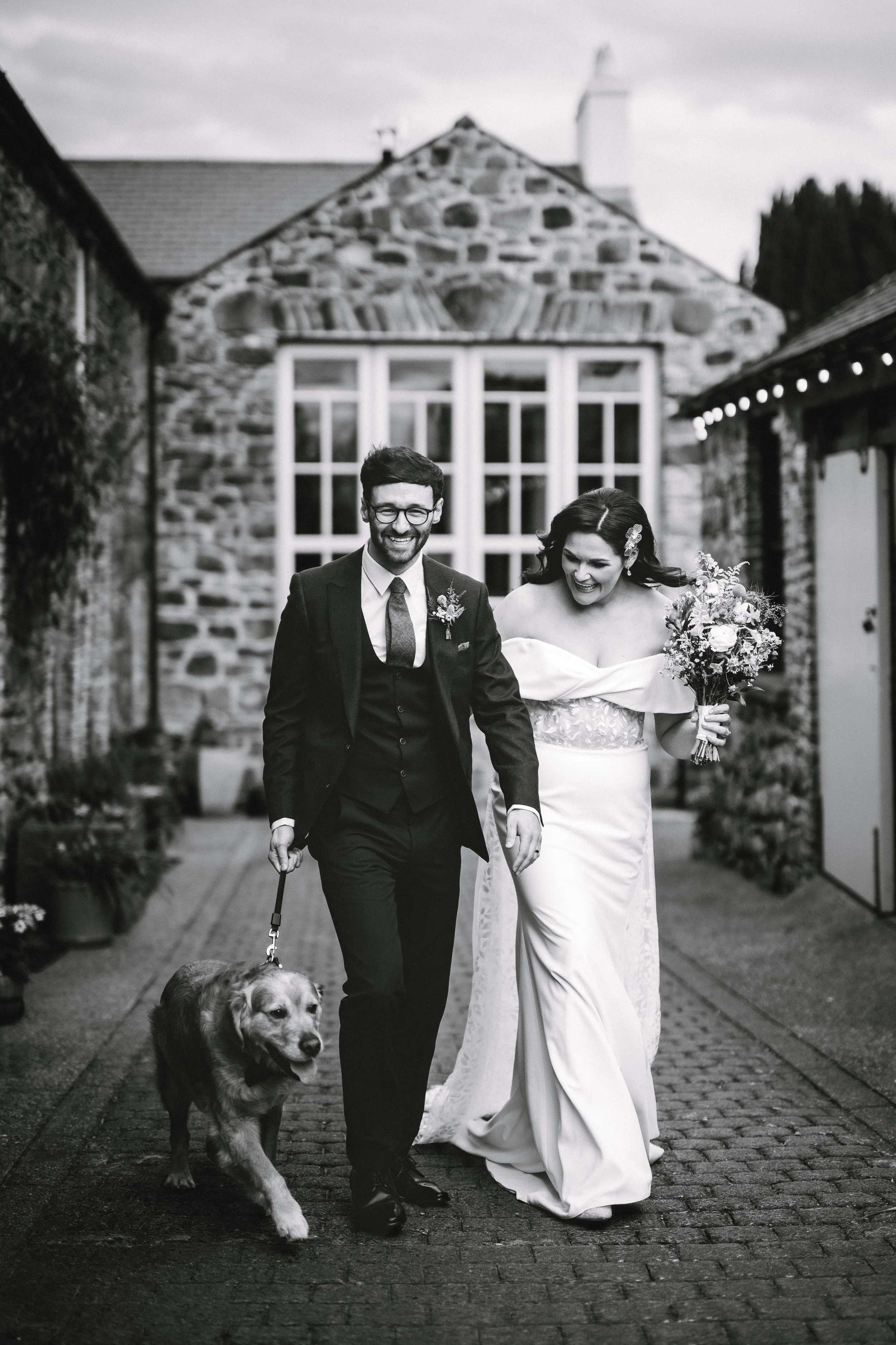 Black and white photo of a happy bride and groom walking with their dog outside a stone house, wedding celebration.