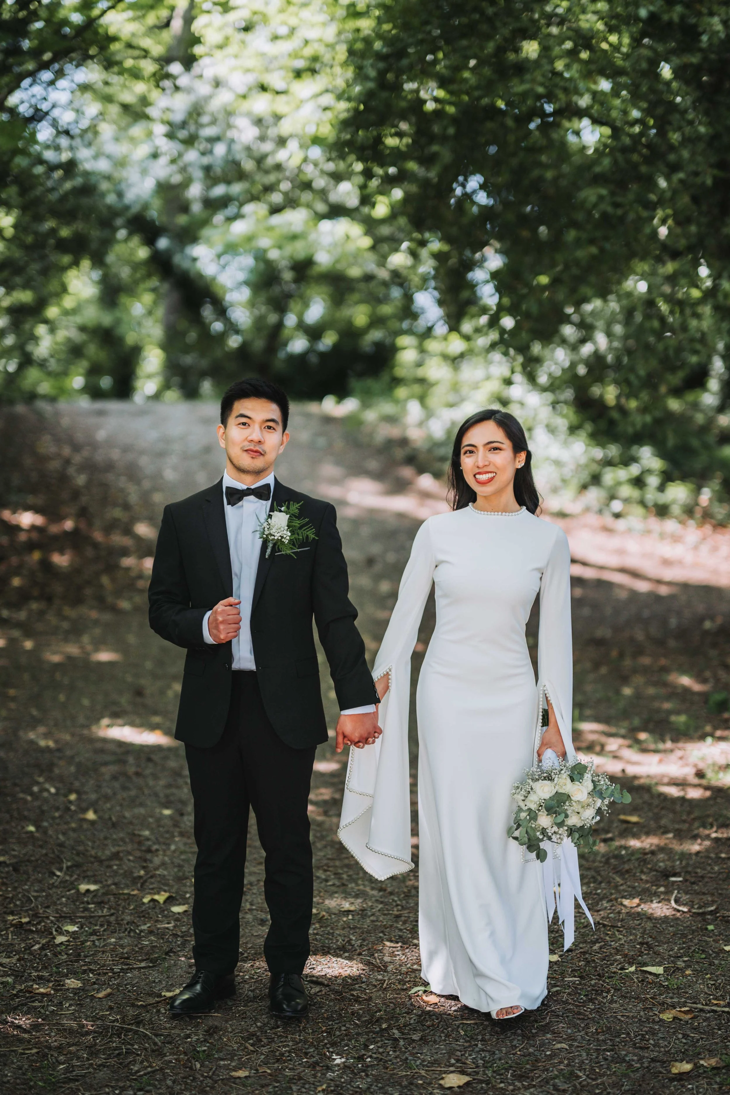 A newlywed couple holding hands in a park, with the groom in a black tuxedo and bow tie, and the bride in a long white dress holding a bouquet of white flowers and greenery.