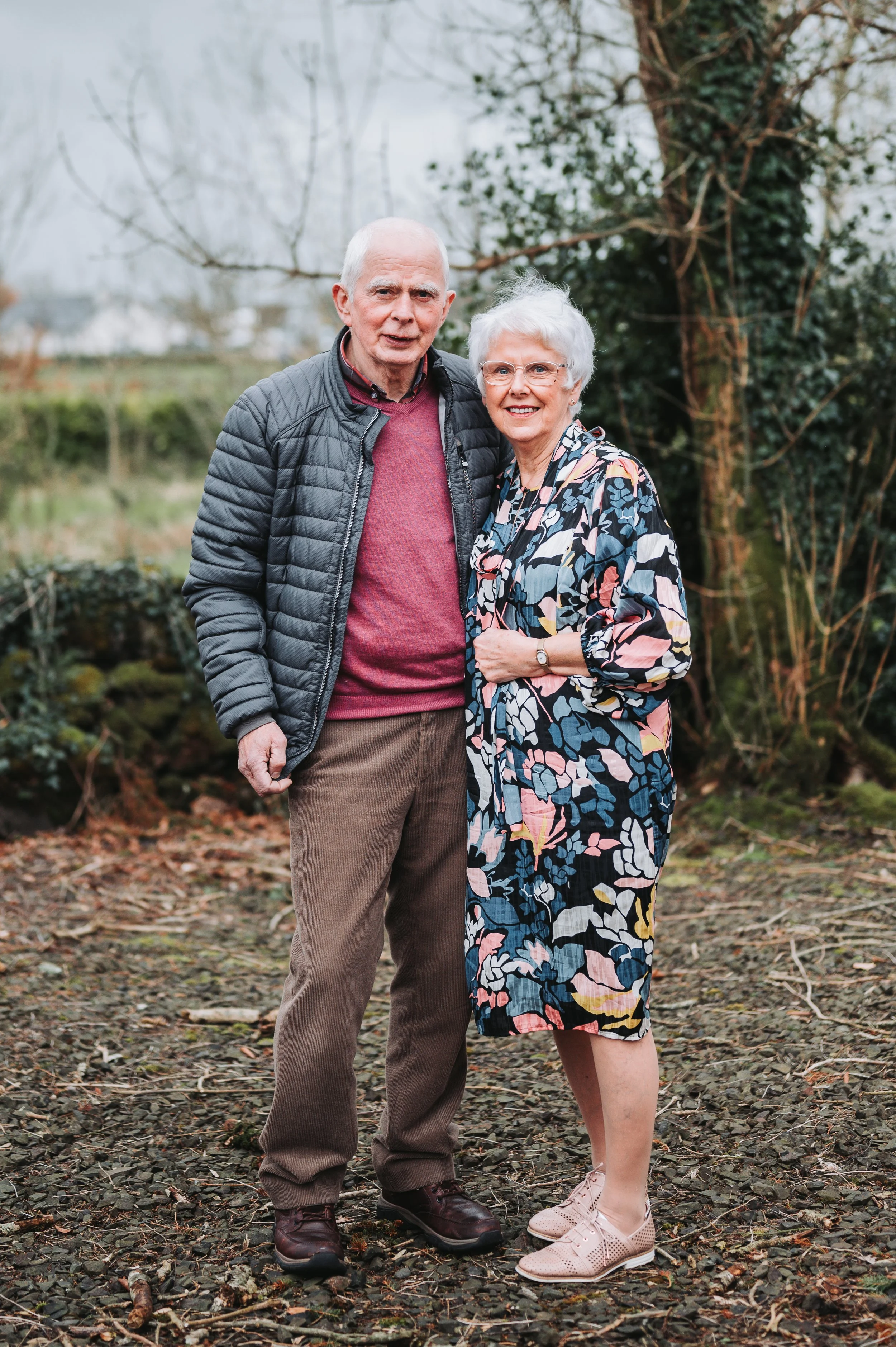 An elderly man and woman standing outdoors on a dirt and gravel path, smiling at the camera, with trees and bushes in the background.
