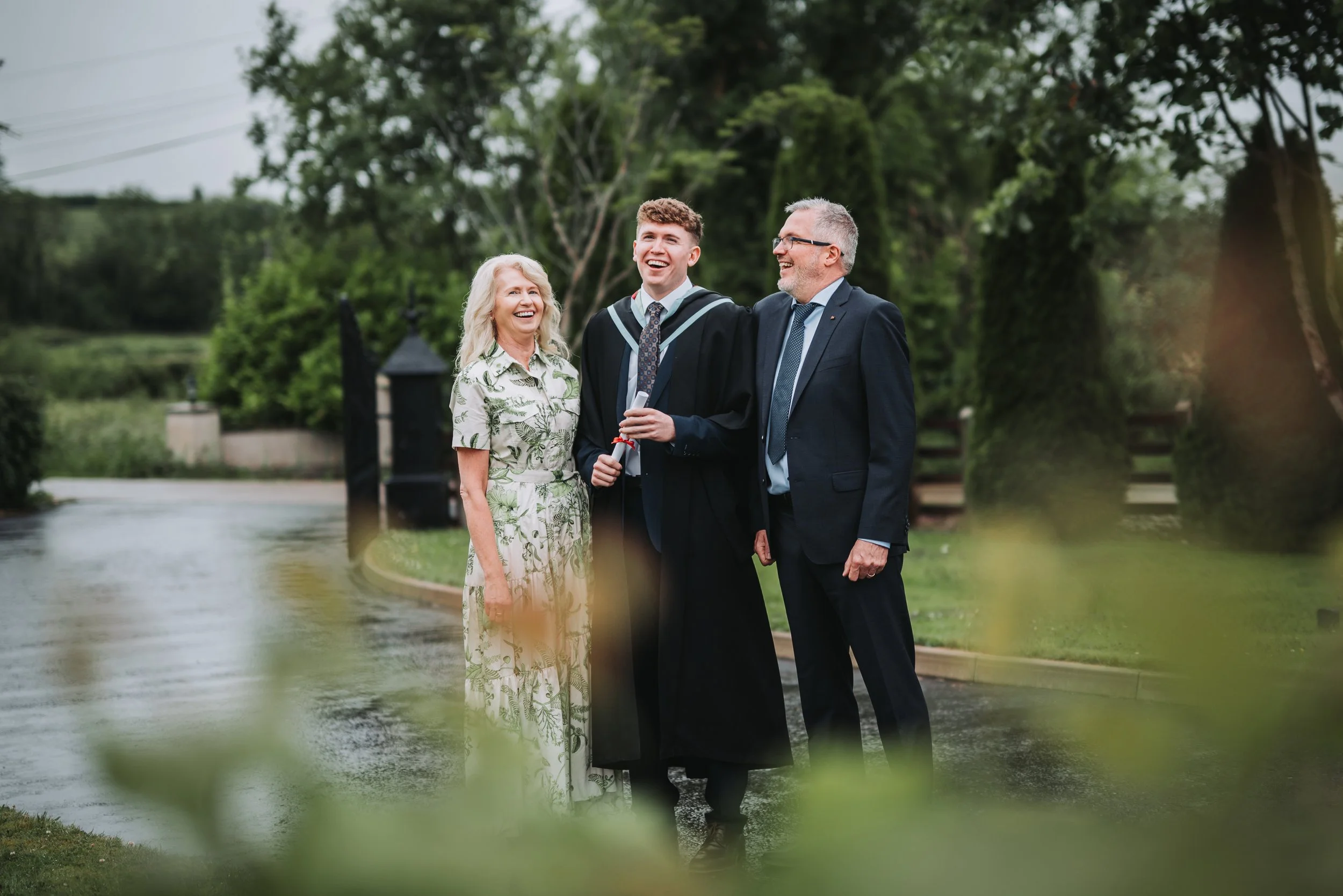 A young man in a graduation gown holding a diploma, smiling with parents outdoors.