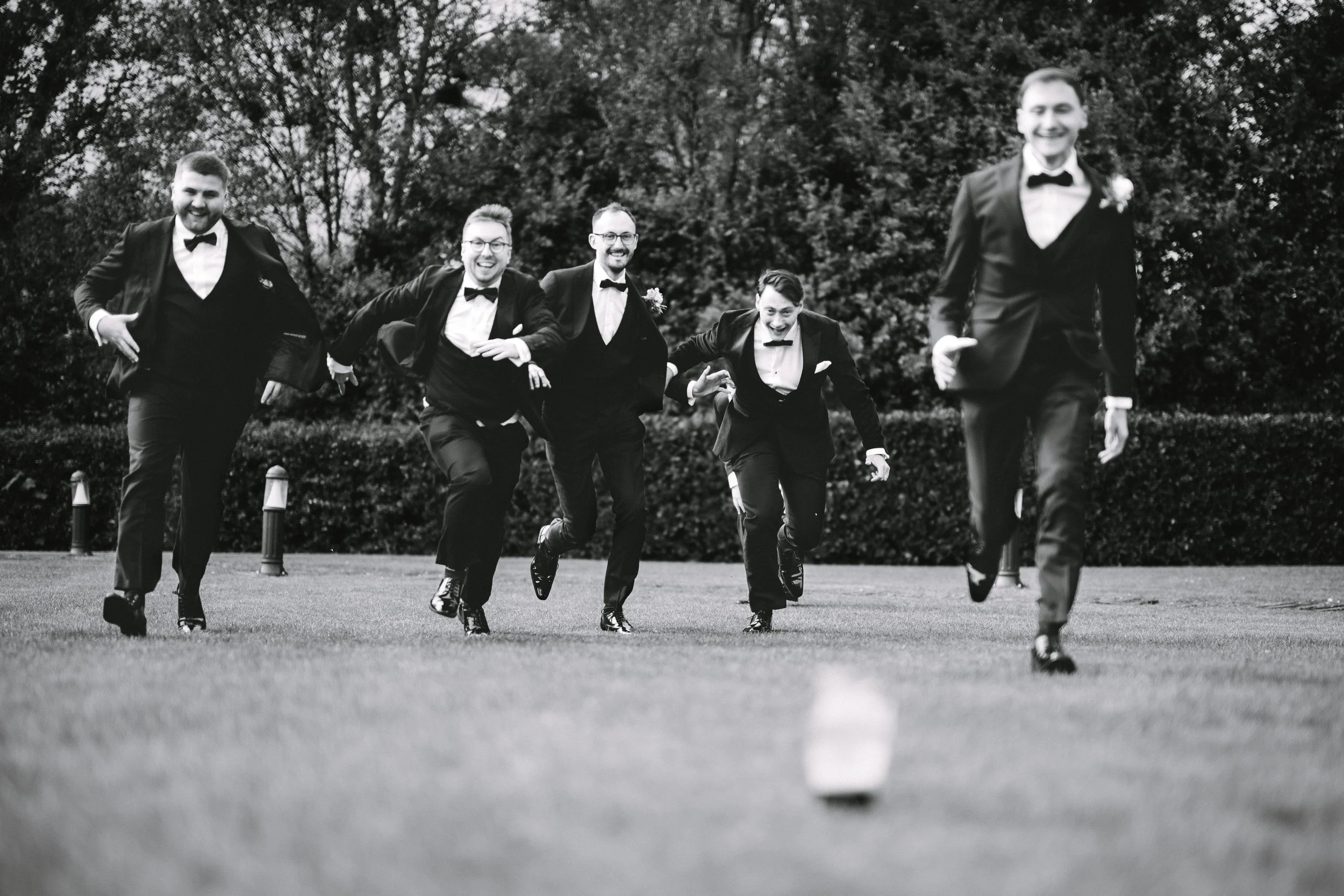 Group of men in tuxedos running towards the camera in an outdoor setting, smiling and laughing.