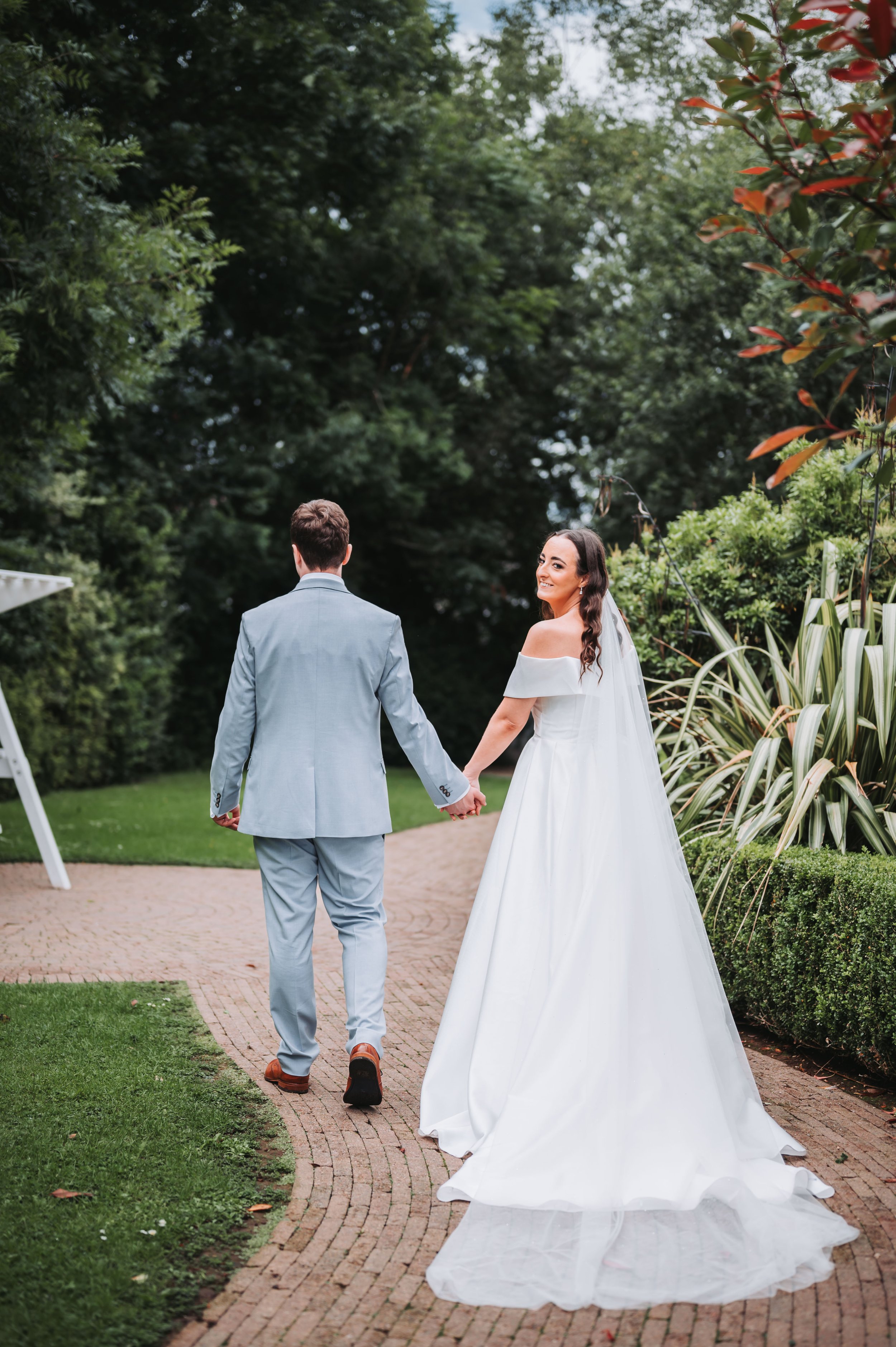 A bride and groom holding hands and walking on a garden path during their wedding, with lush green trees and bushes surrounding them.