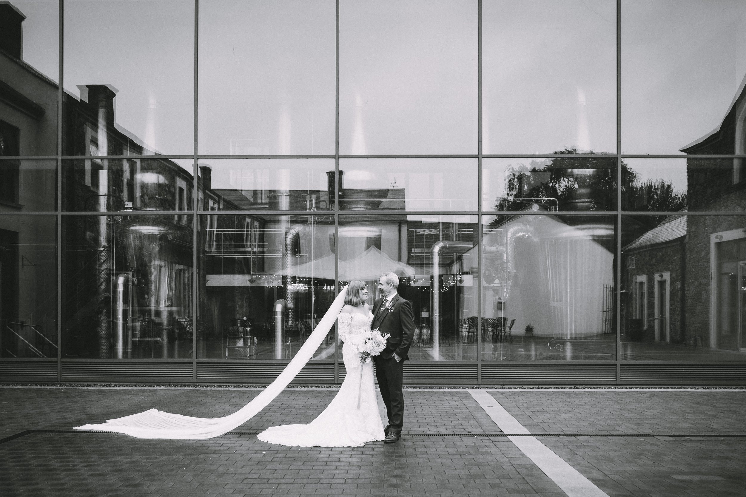 Black and white photo of a bride and groom standing outside a modern building with large glass windows, reflection of surrounding buildings visible, bride in a wedding gown with long train, holding a bouquet, groom in a suit.