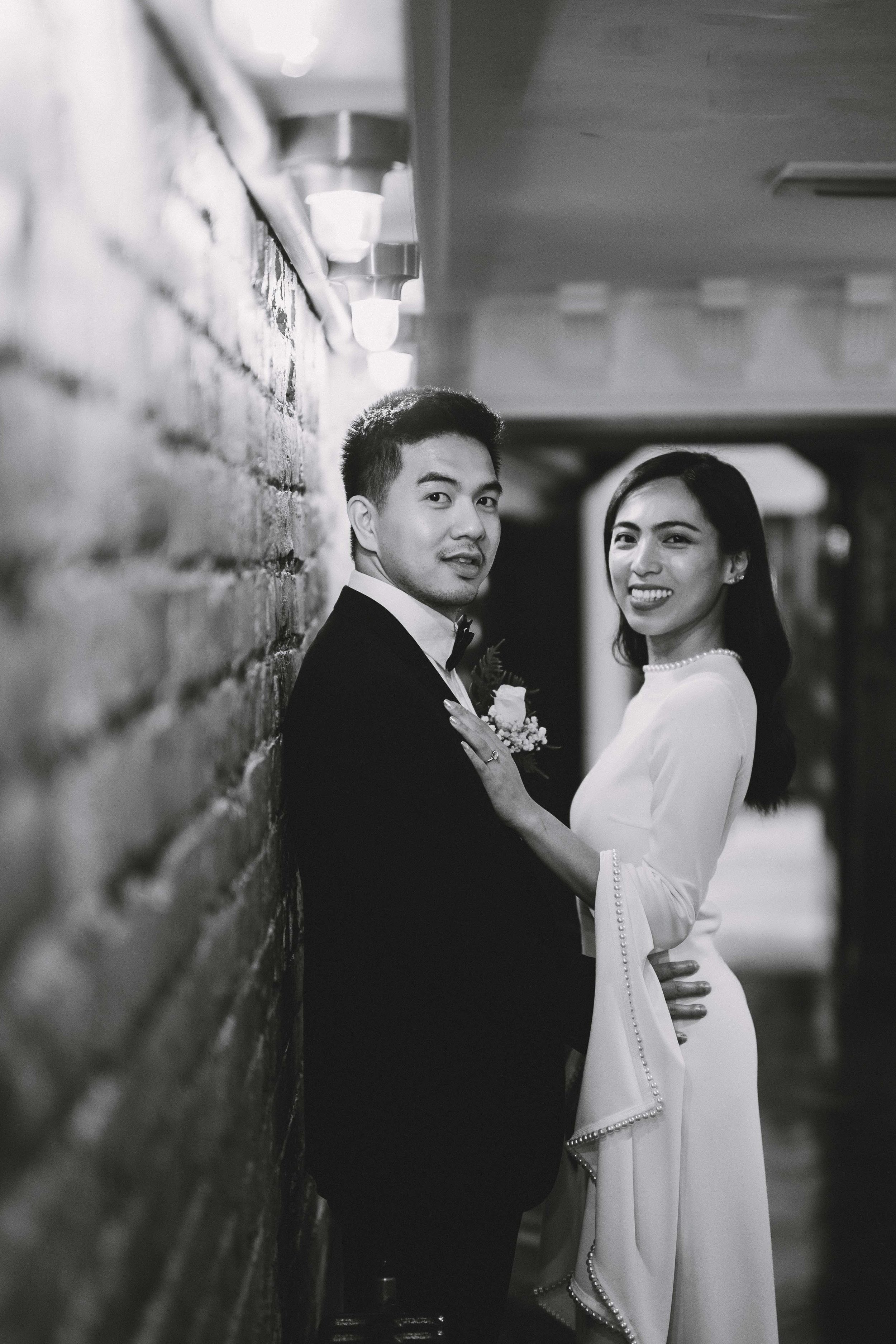 Black and white photo of a happy couple in wedding attire, standing close to each other, smiling, with the woman touching the man's chest, against a textured brick wall in an indoor setting.
