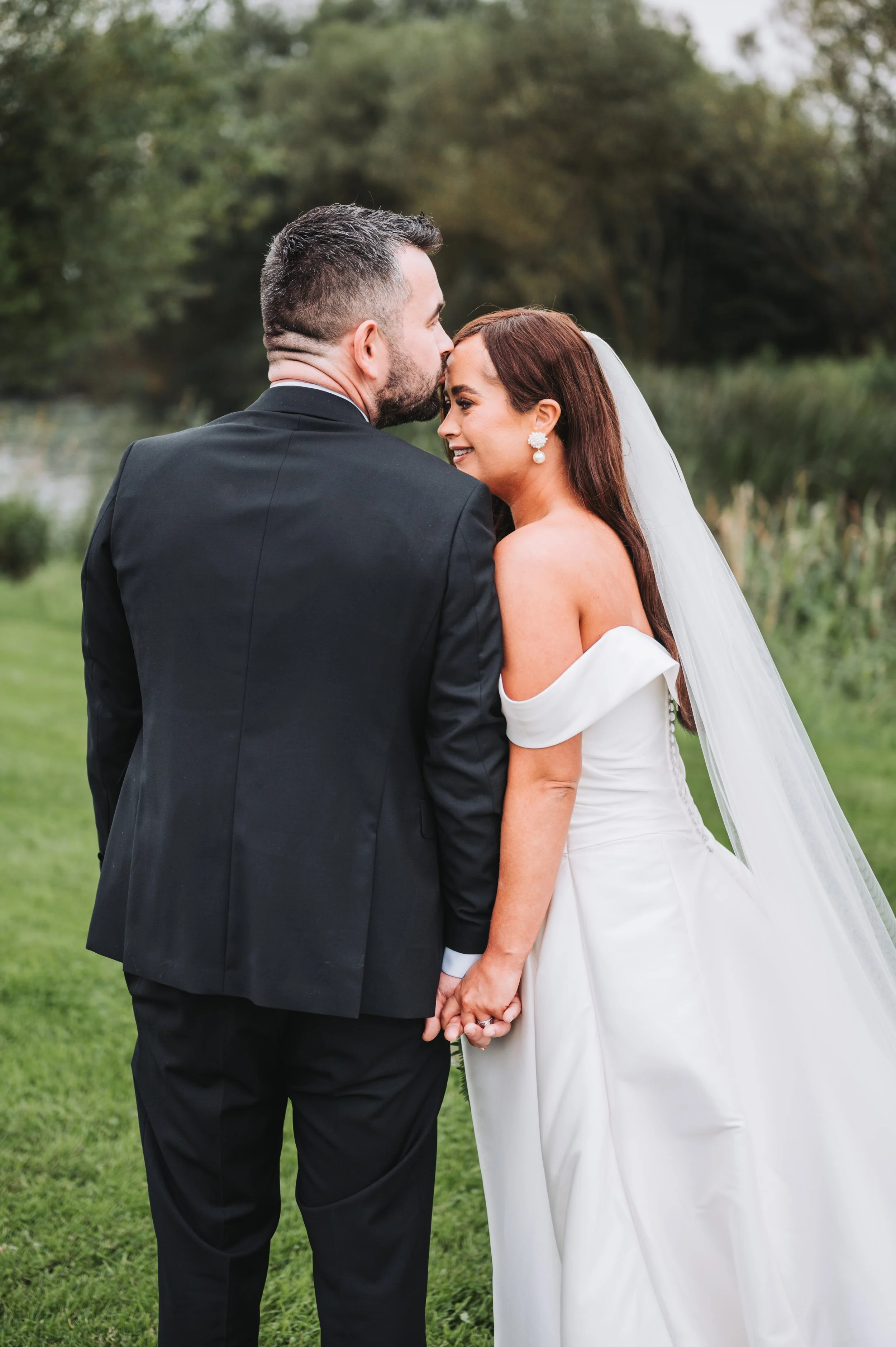 A newlywed couple holding hands in an outdoor setting, with the groom kissing the bride's forehead during daytime.