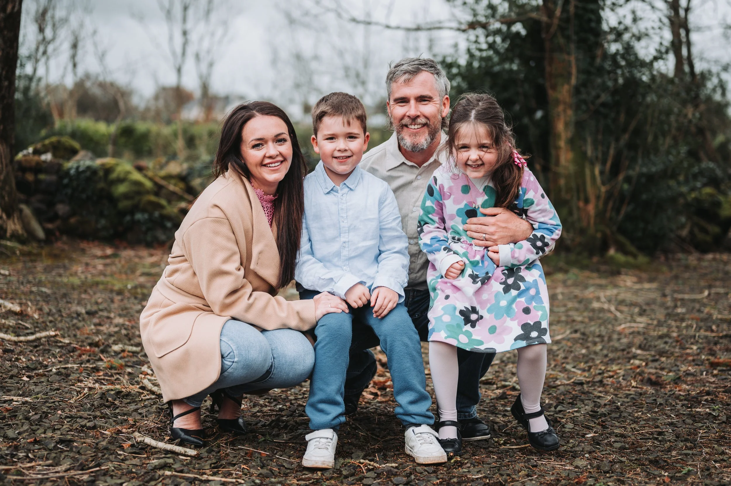 A smiling family of four posing outdoors in a wooded area, with two parents and two children, a boy and a girl, all dressed casually.