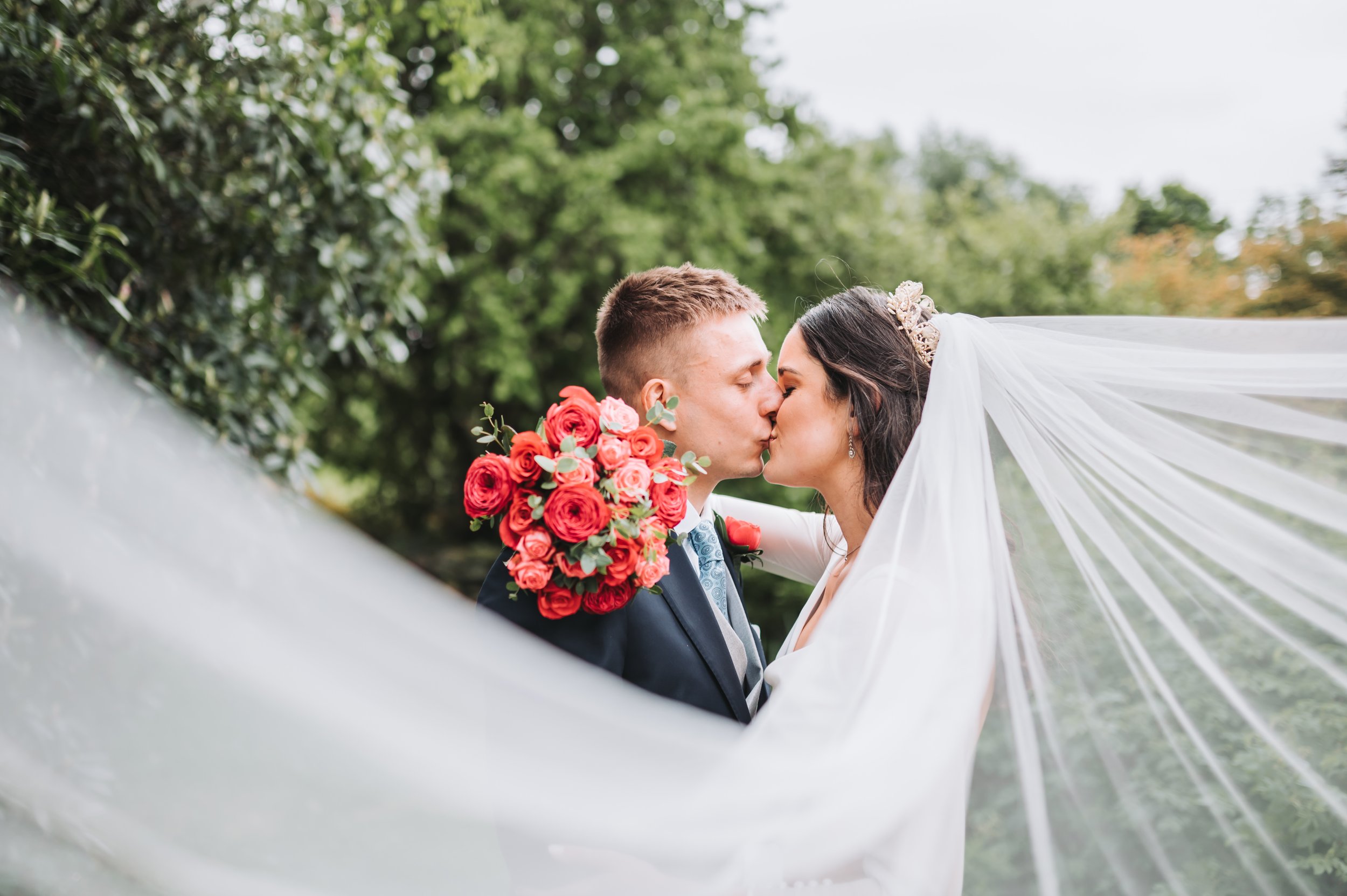 Bride and groom kissing with bouquet and flowing veil, outdoor wedding setting.