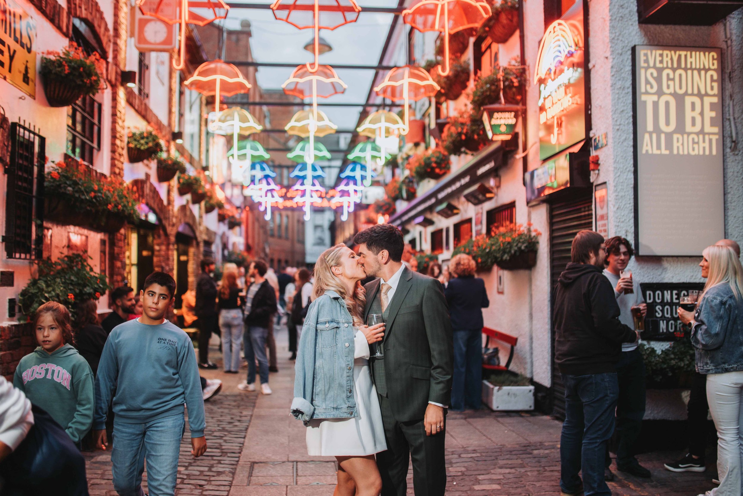 A couple kisses on a lively city street decorated with colorful umbrella neon lights, with multiple people socializing and walking around.