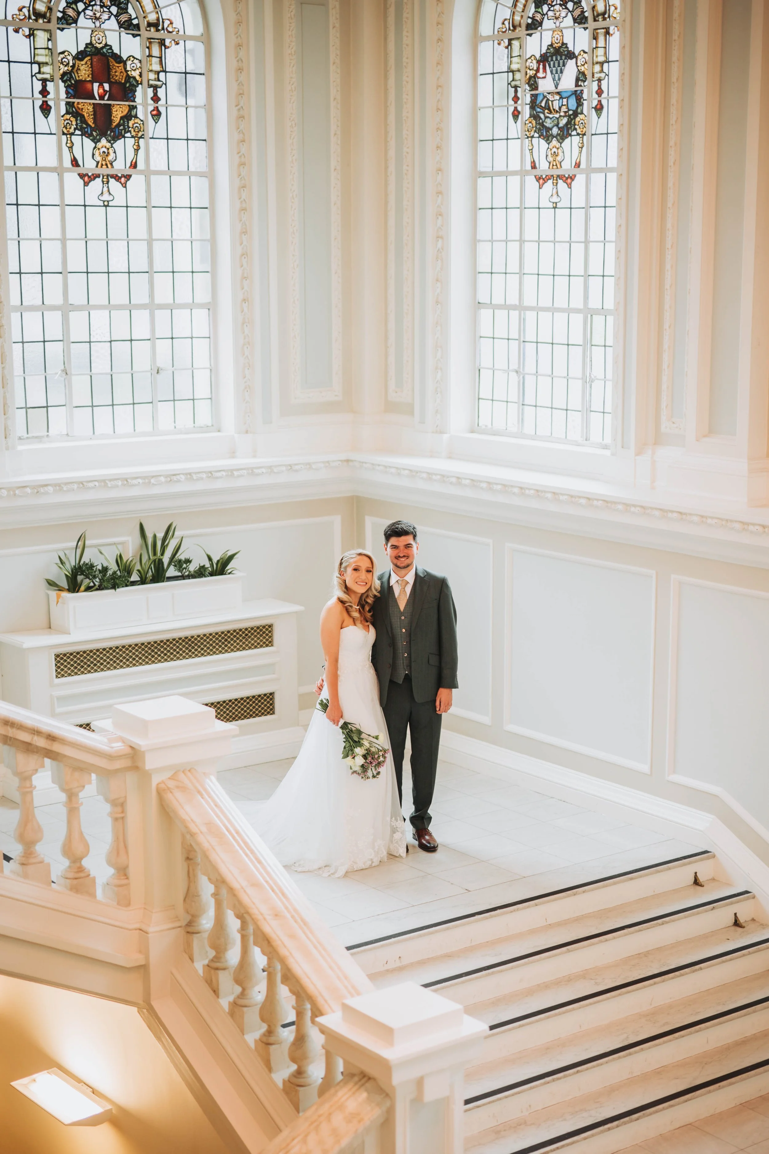 A bride and groom standing together at the top of a staircase inside a bright, elegant building with large stained glass windows.
