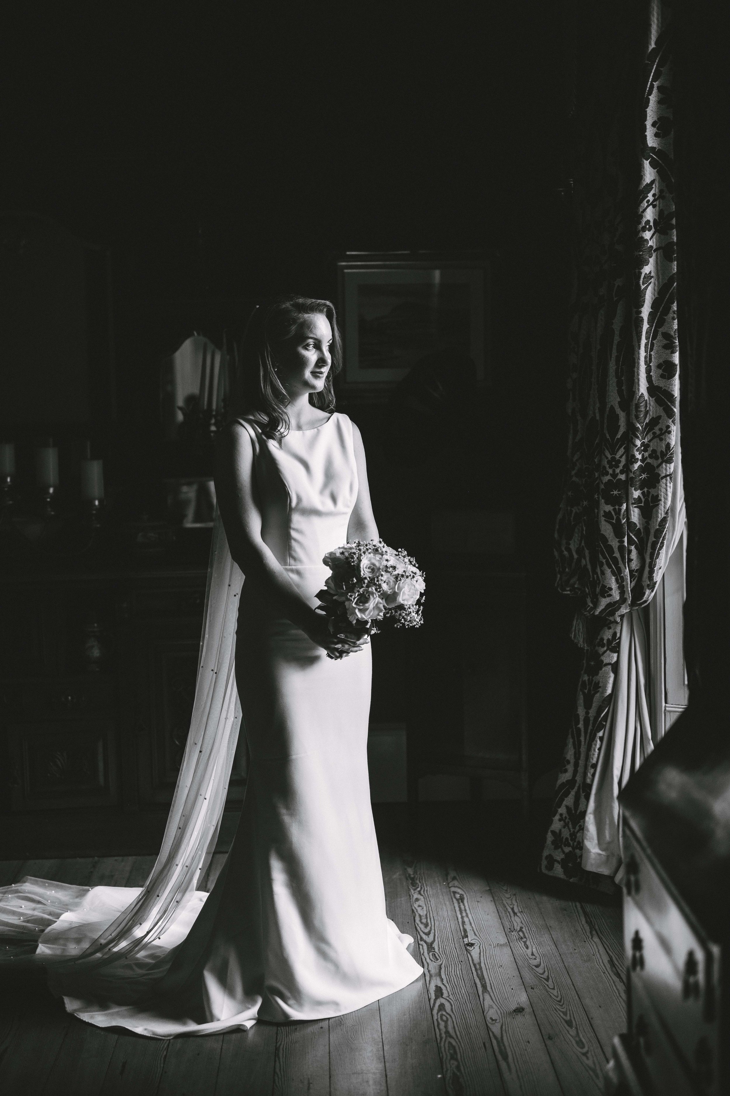 A bride in a sleeveless wedding gown holding a bouquet of flowers, standing by a window with patterned curtains, looking thoughtfully outside in a dimly lit room.