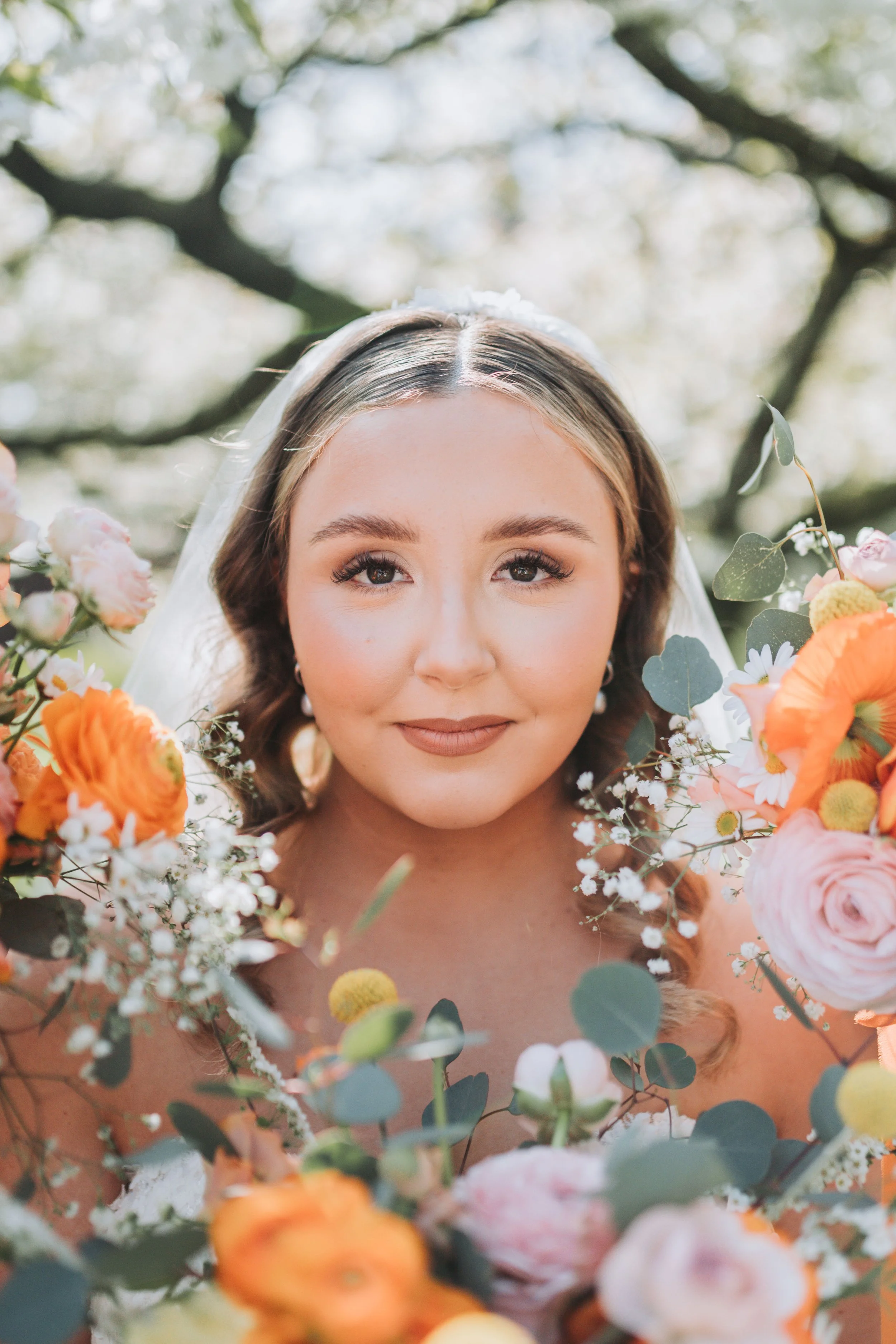 Close-up of a bride with light makeup and long wavy hair, surrounded by colorful flowers including roses and daisies, outdoors with tree branches in the background.