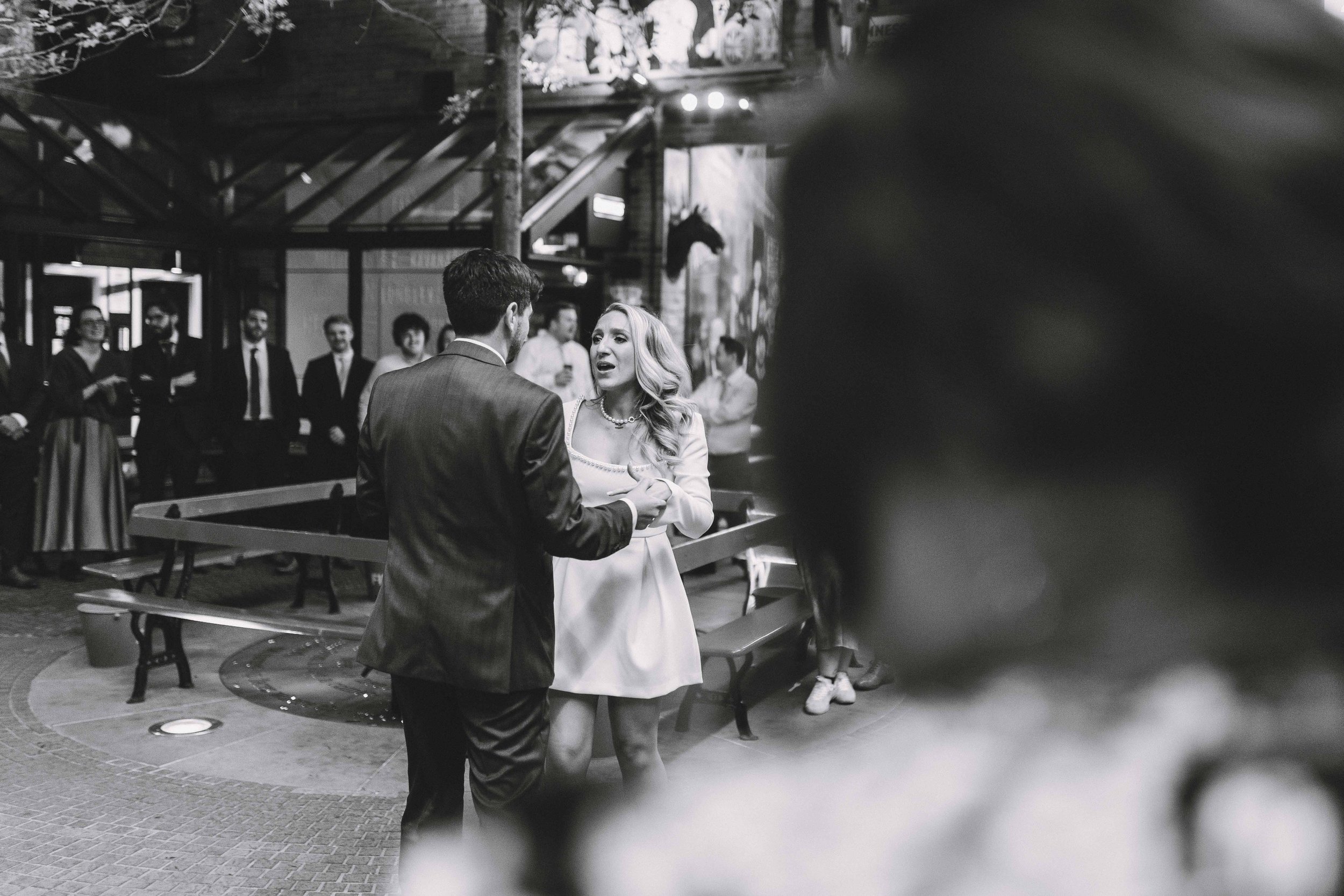 A couple dancing at a wedding reception, with guests in the background, in black and white.