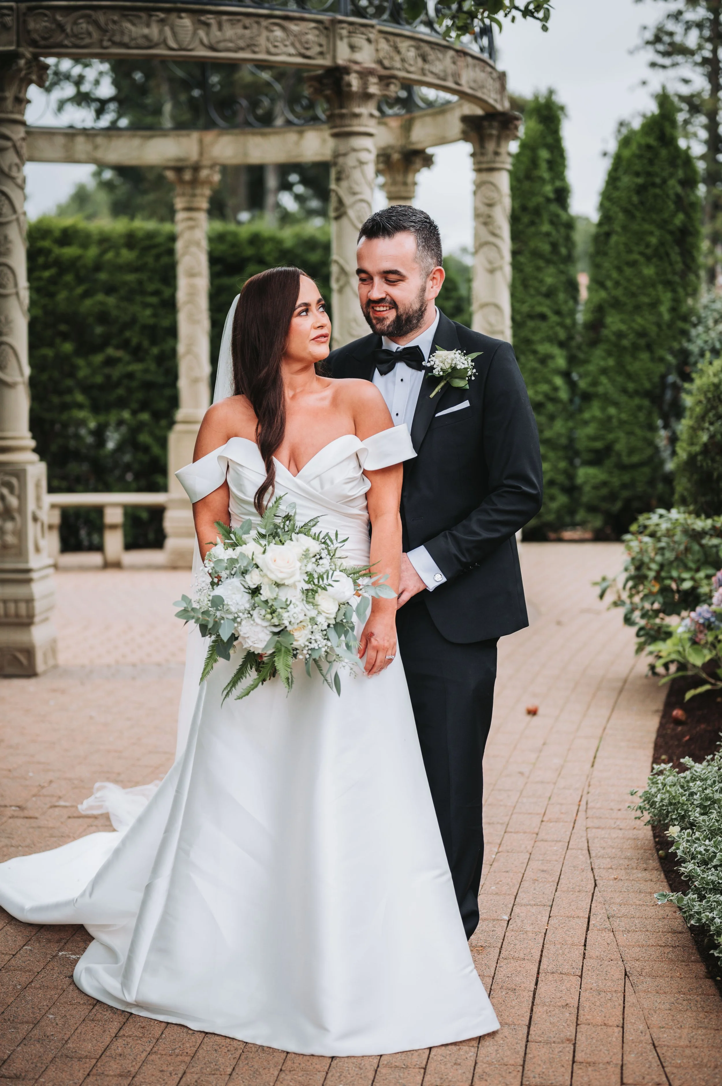 A bride in an off-shoulder white wedding gown holding a bouquet of white roses and greenery, standing close to a groom in a black tuxedo with a black bow tie, outdoors with an ornate stone gazebo and greenery in the background.