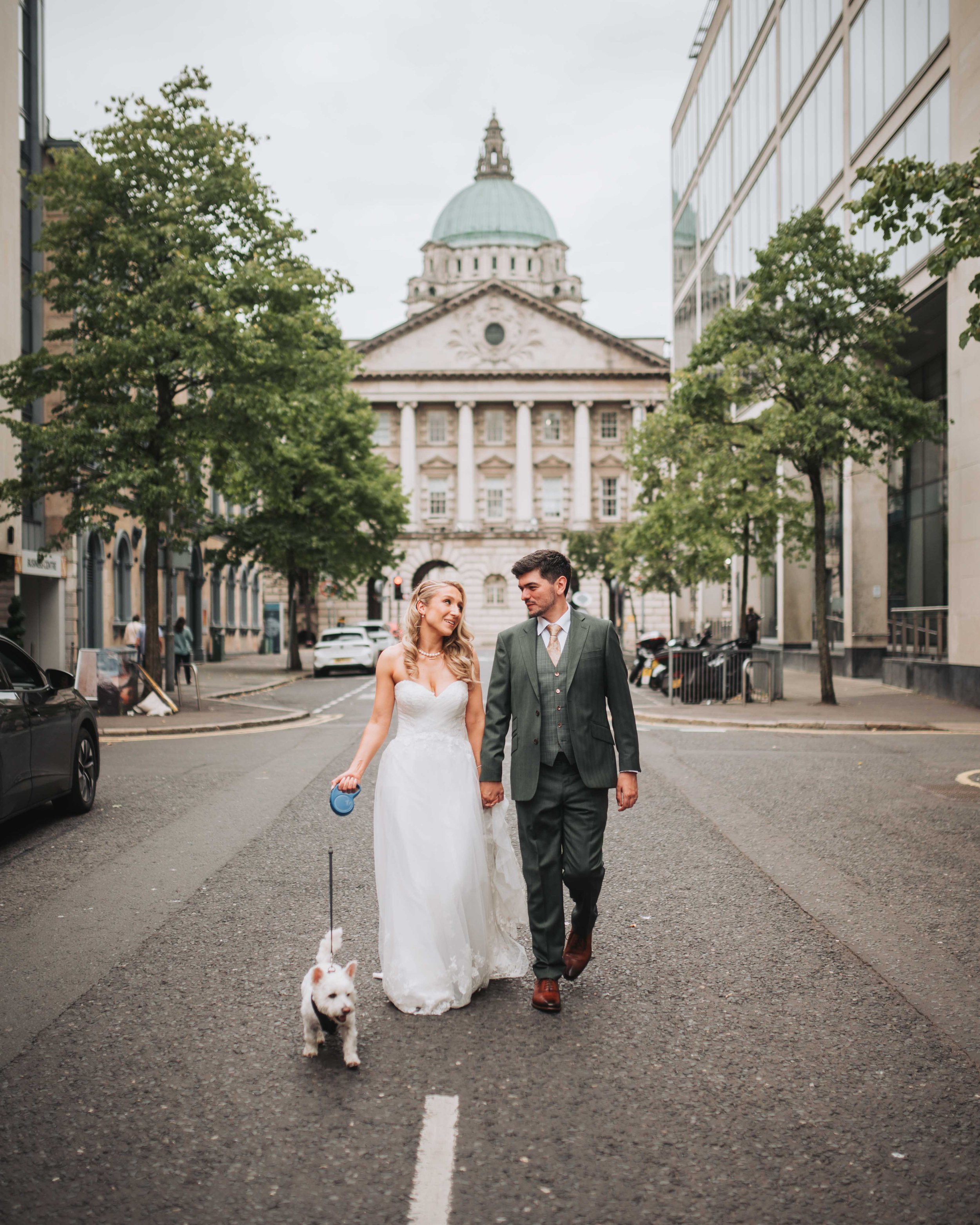 A bride and groom walking hand in hand on a city street with a small white dog, in front of historic building with a dome, surrounded by trees and modern buildings.