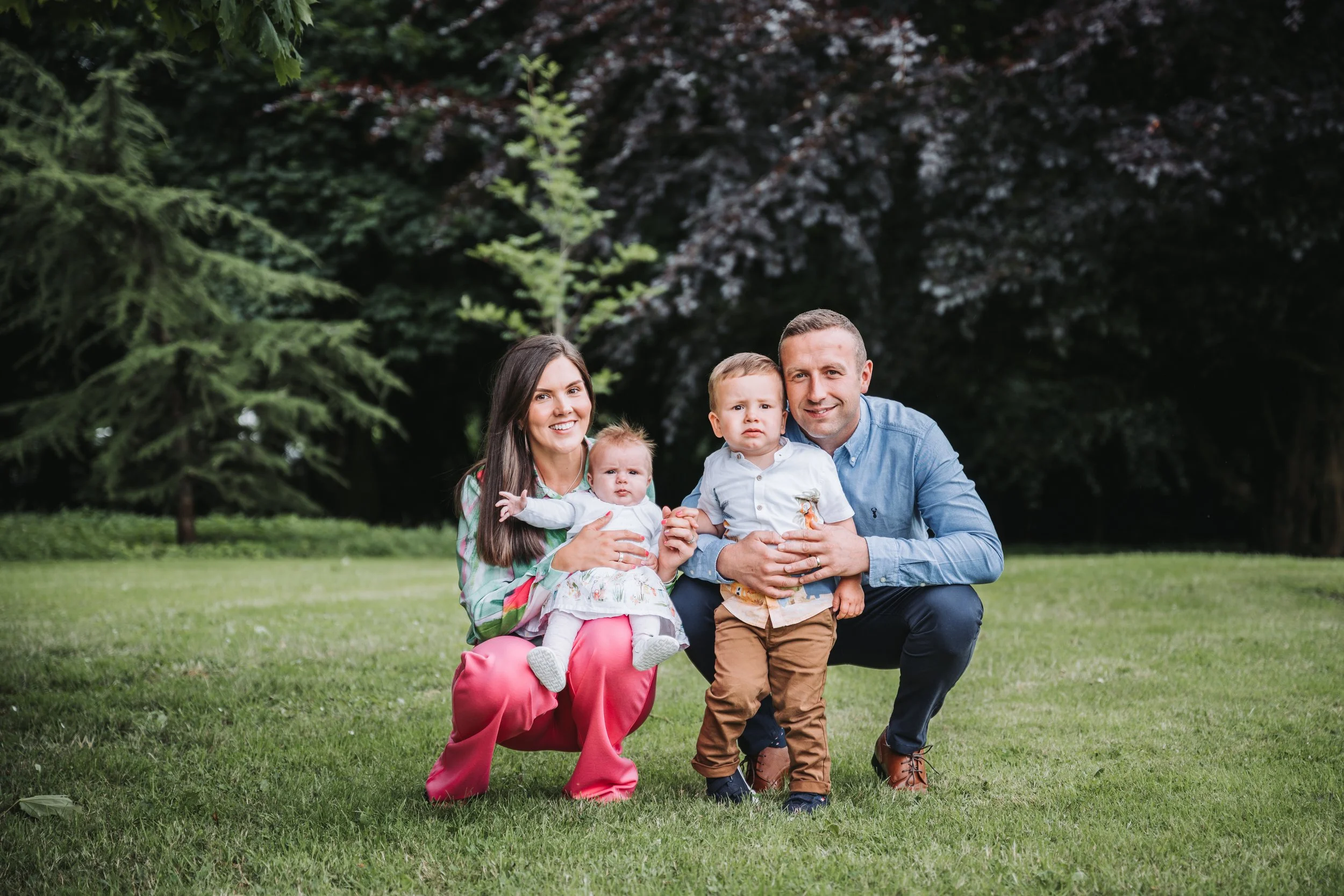 Family of four with a woman, man, and two young children, posing outdoors on grass with trees in the background.