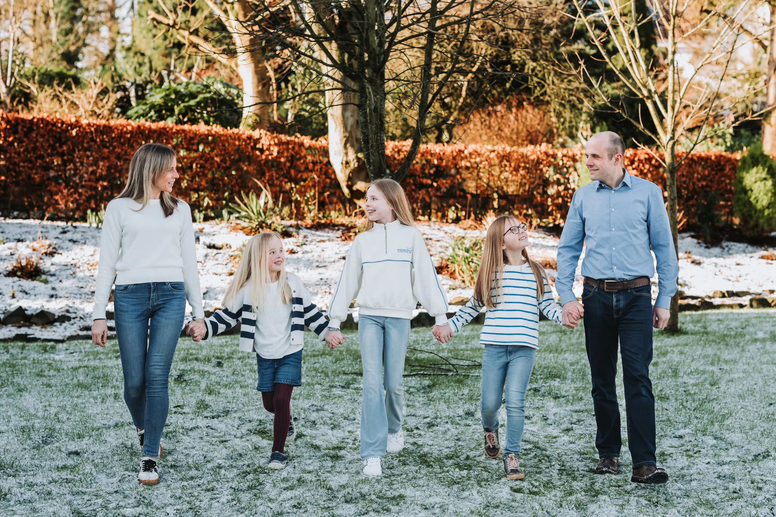 A family of six, including two adults and four children, walking hand in hand outdoors on a grassy area with some light snow, trees, and bushes in the background during autumn.