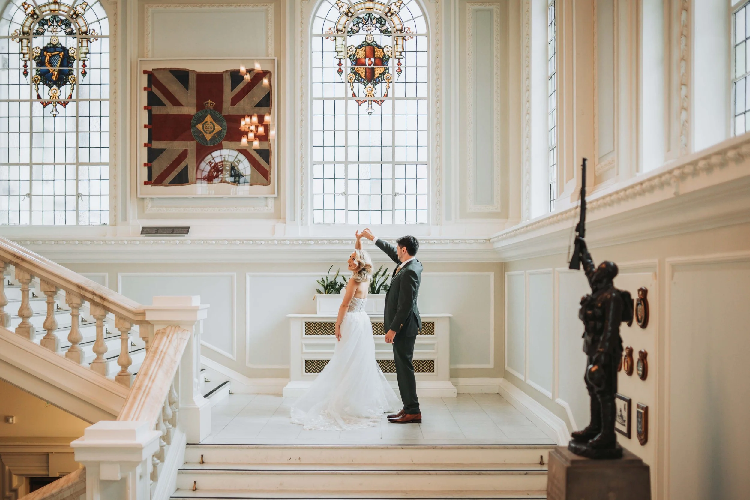 A bride and groom dancing in a grand, well-lit hall with large stained-glass windows and tall, ornate ceilings. The bride is in a white wedding dress, and the groom is in a dark suit. There is a British flag painting on the wall behind them, and a so