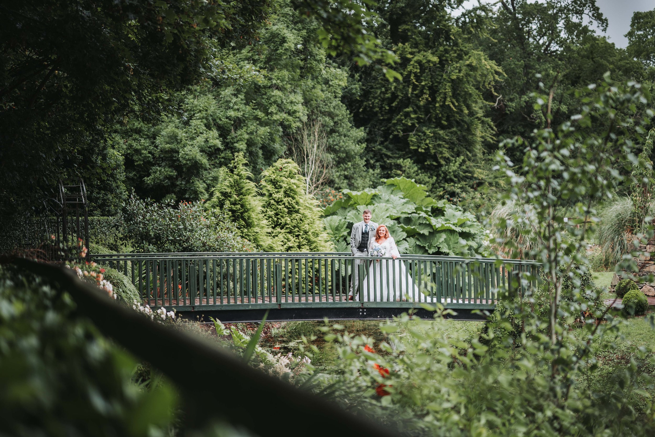 A bride and groom standing on a bridge in a lush garden, surrounded by green trees and plants, with the bride in a white wedding dress holding a bouquet.