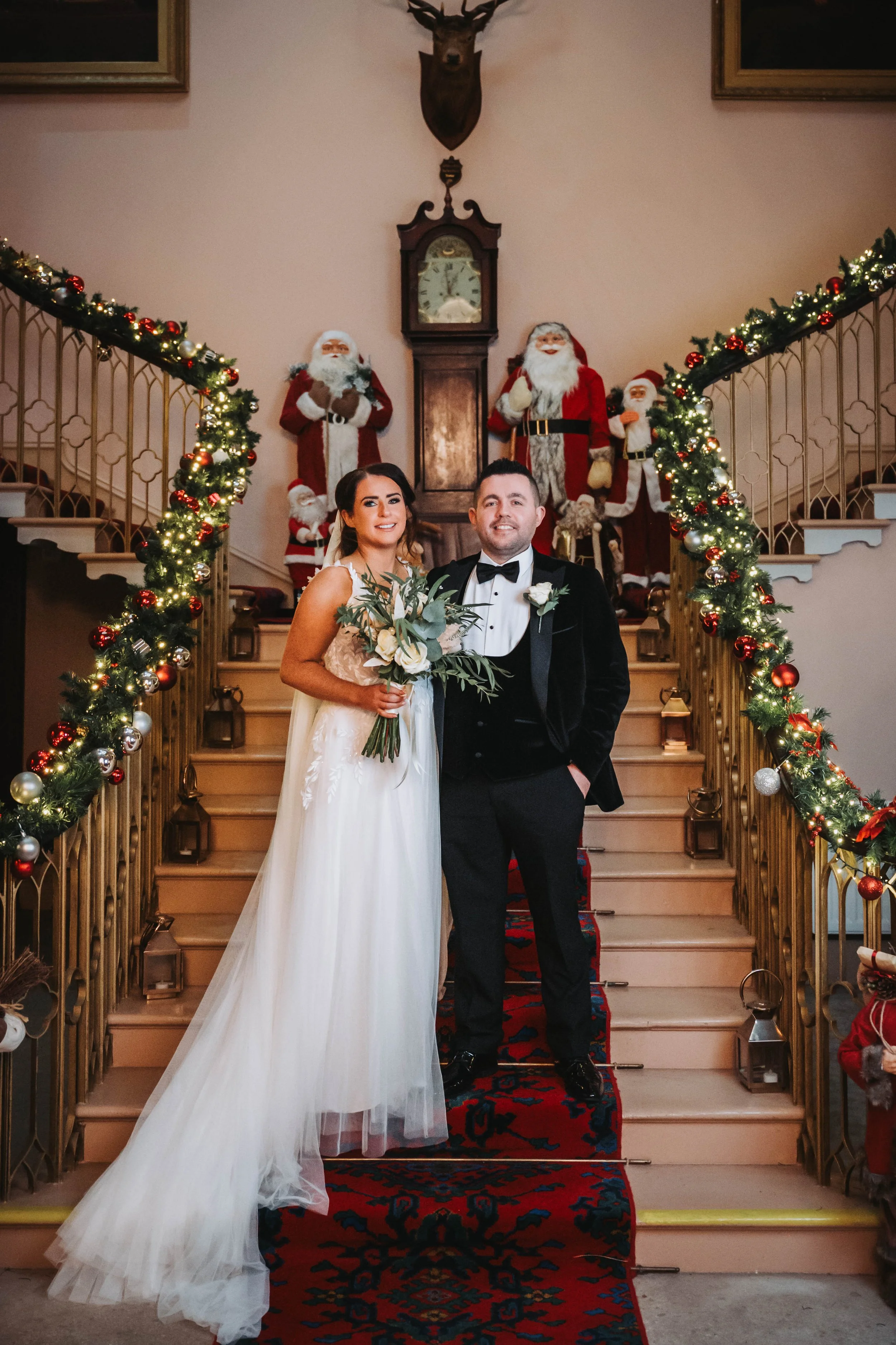 A bride and groom standing on a staircase decorated with Christmas garlands, posing for a wedding photo with Christmas-themed Santa Claus figures and a vintage clock in the background.