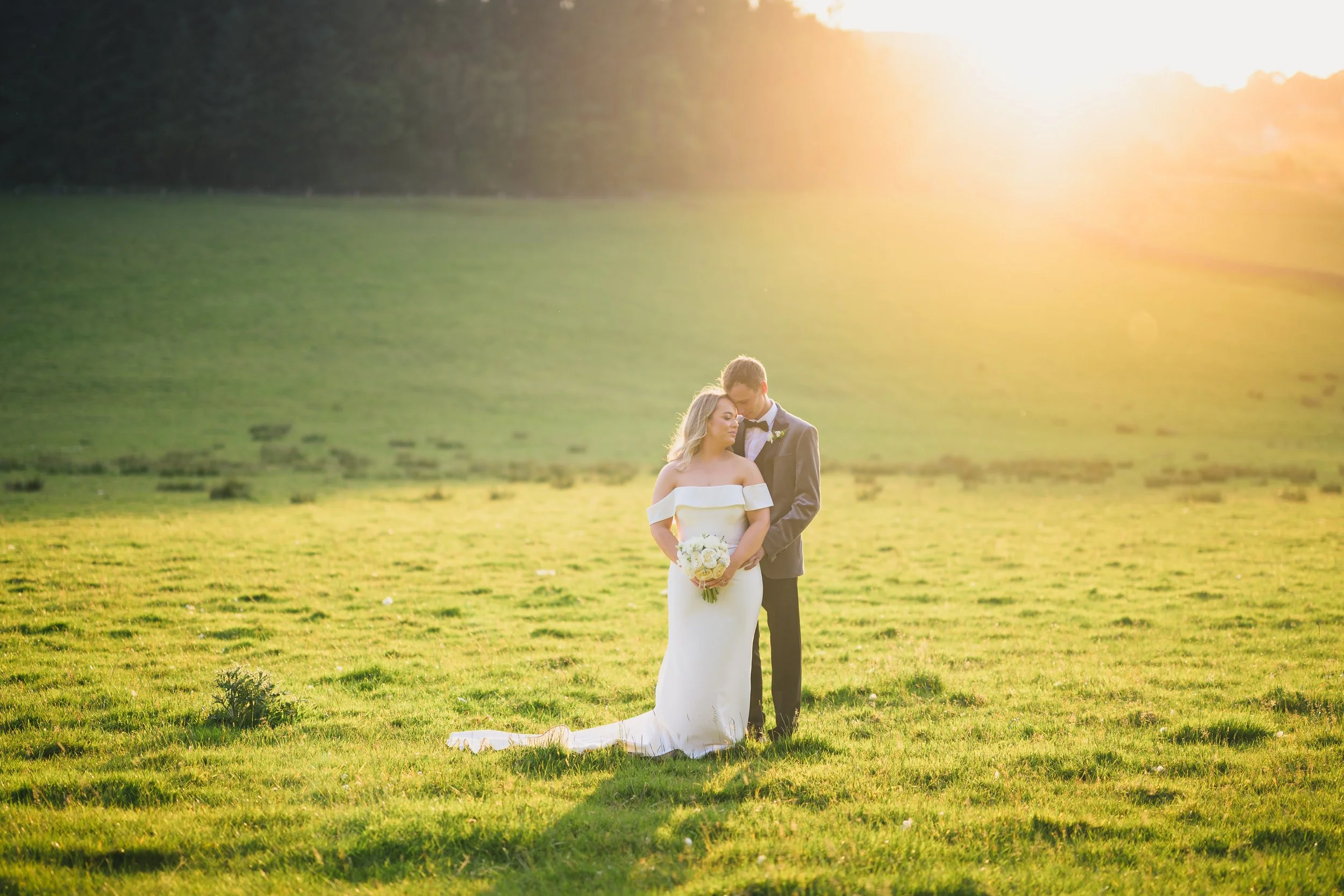 A bride in a white off-shoulder wedding dress and a groom in a black tuxedo standing closely in a green field during sunset, with the bride holding a bouquet of white flowers.