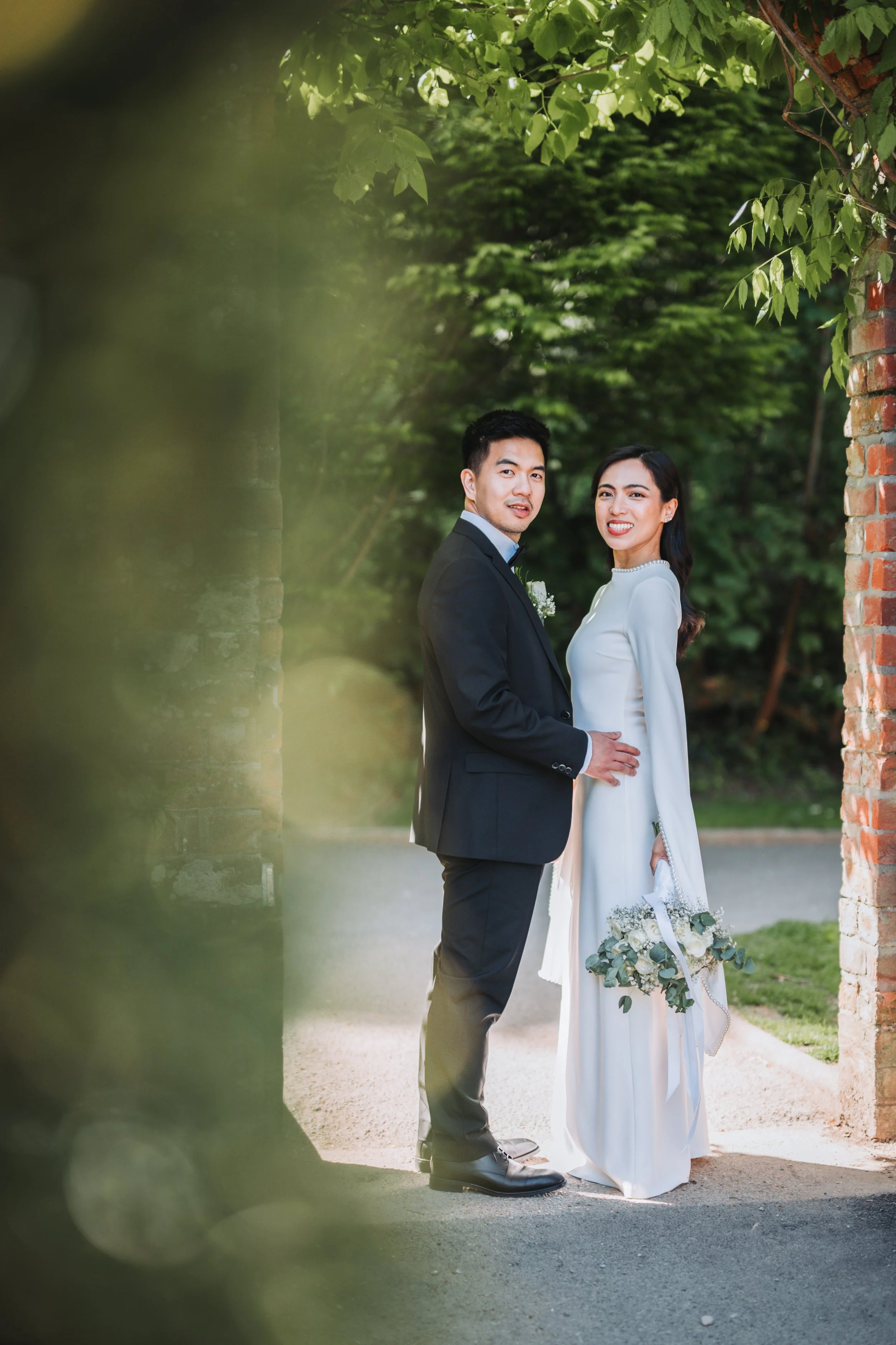 A newlywed couple standing outdoors near a rustic brick wall, with lush green trees in the background; the groom is in a black suit and the bride is in a white dress holding a bouquet of flowers.