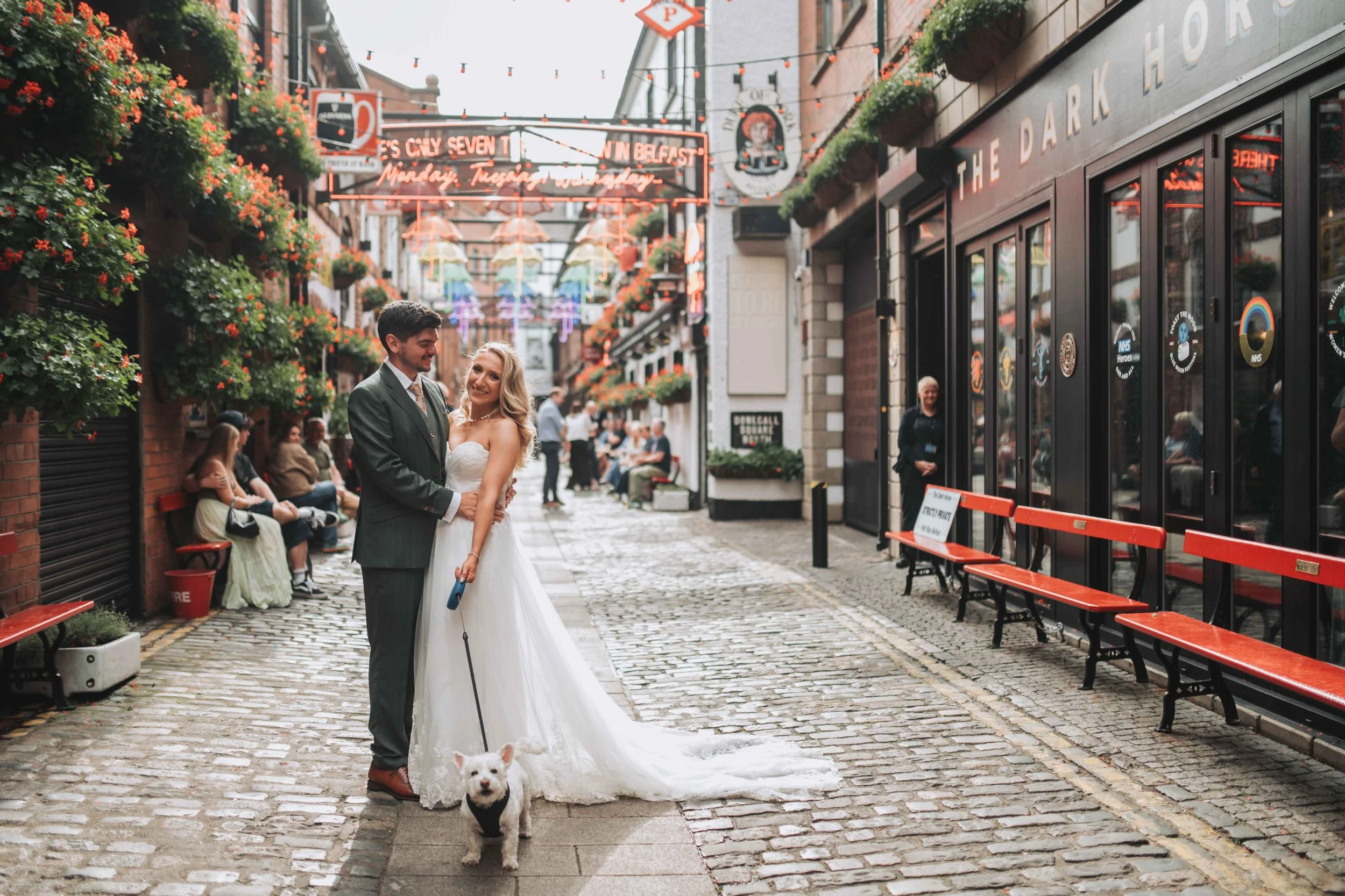 A bride and groom standing on a cobblestone street in a city, holding a small white dog, with other people sitting on benches in the background, colorful neon lights hanging above, storefronts on either side, and decorations suggesting a festive atmo