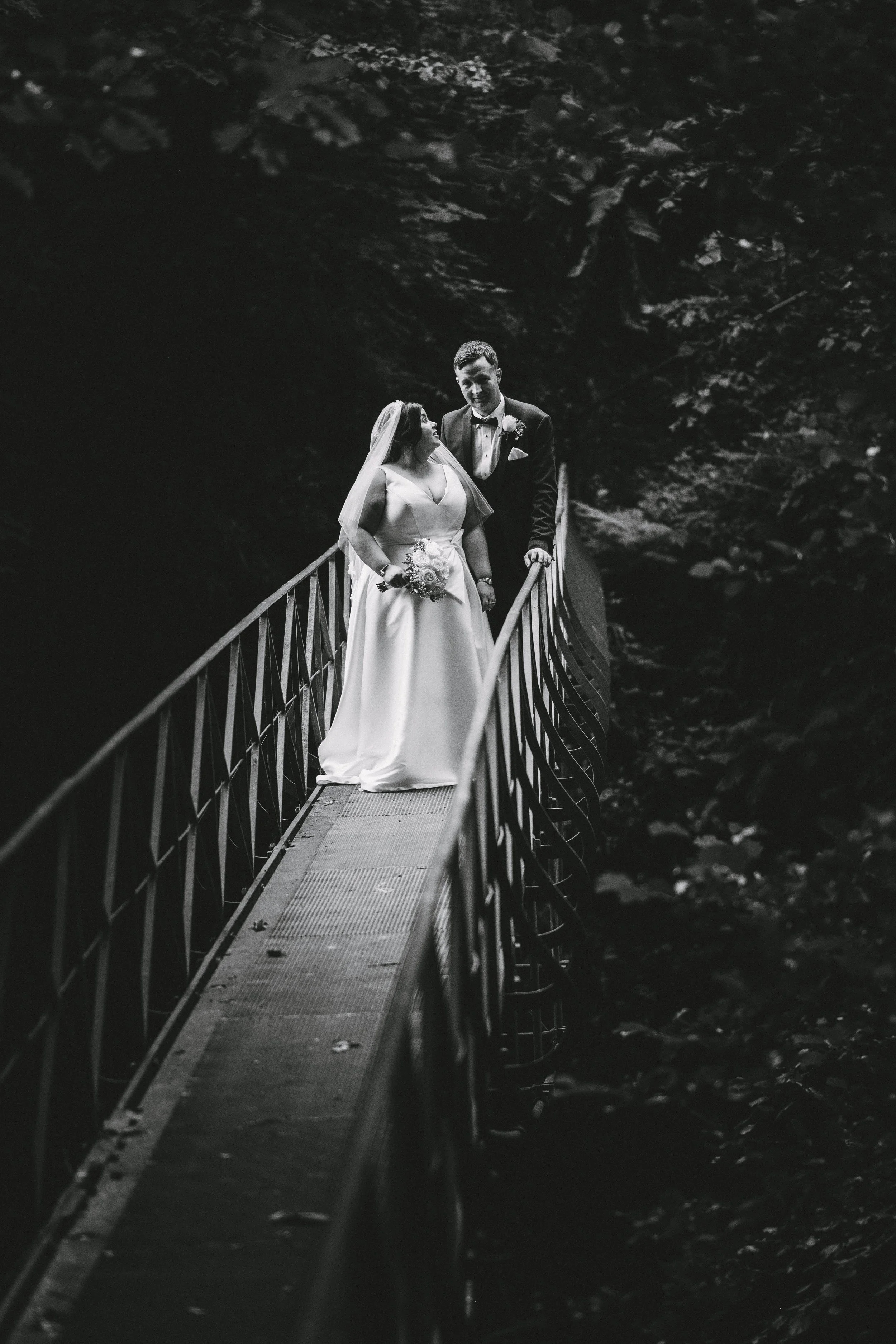 Black and white photograph of a bride and groom standing on a narrow, curved bridge in a wooded area. The bride is holding a bouquet and wearing a wedding dress with a veil, while the groom is in a tuxedo. They are looking at each other.