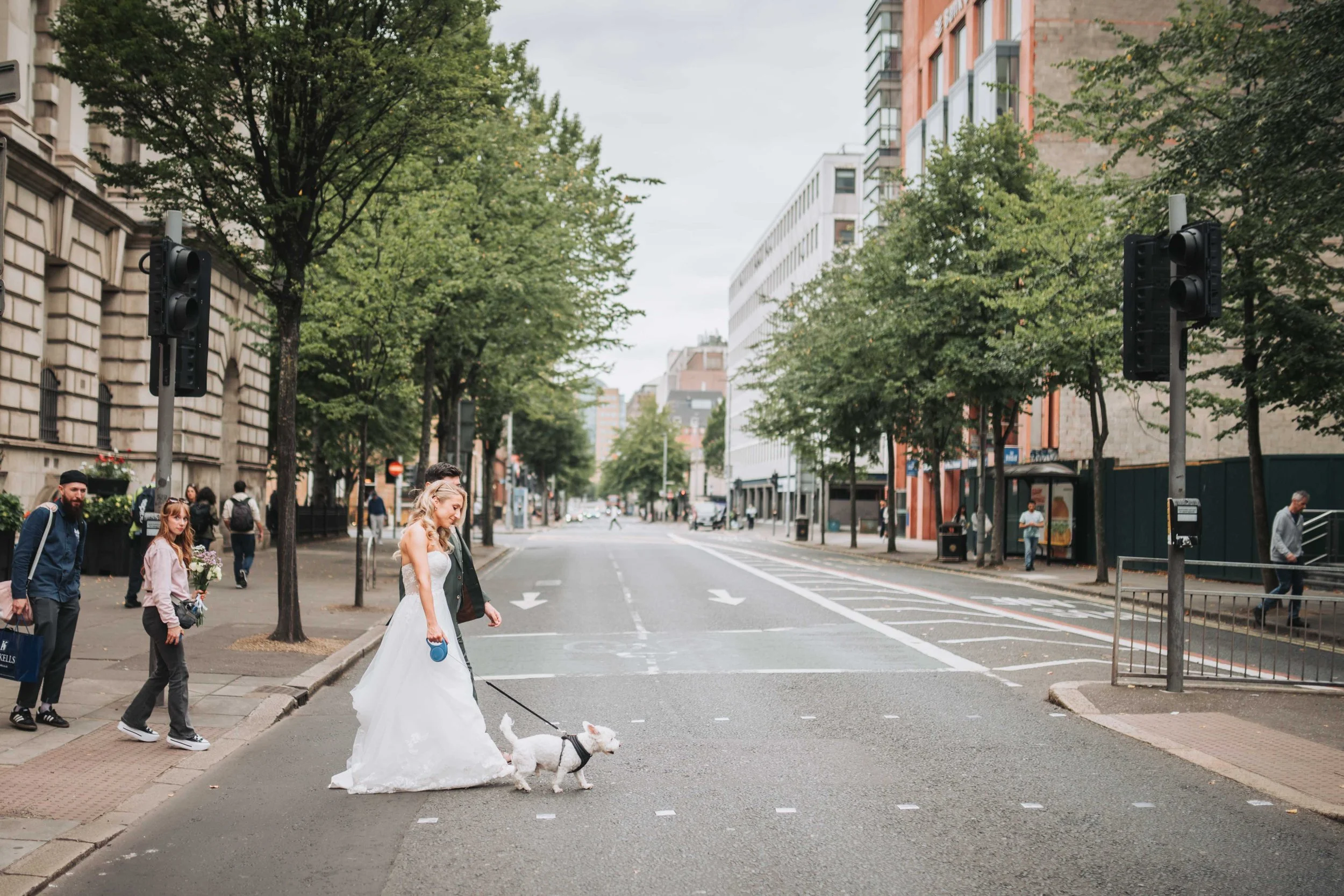 A bride in a white wedding dress walking her dog on a city street while a man walks beside her, with onlookers watching from the sidewalk and traffic lights at an intersection.