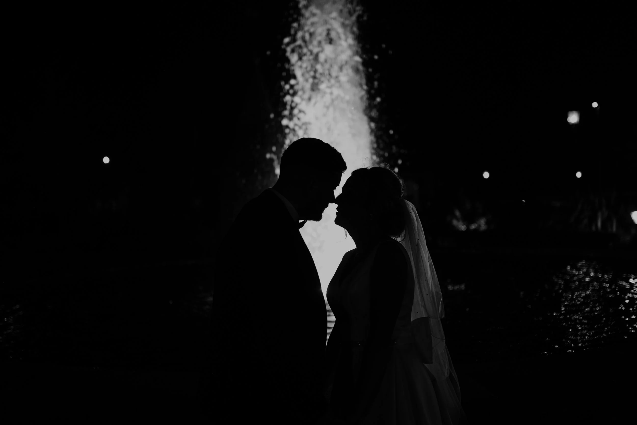 Silhouette of a couple facing each other in front of an illuminated water fountain at night.