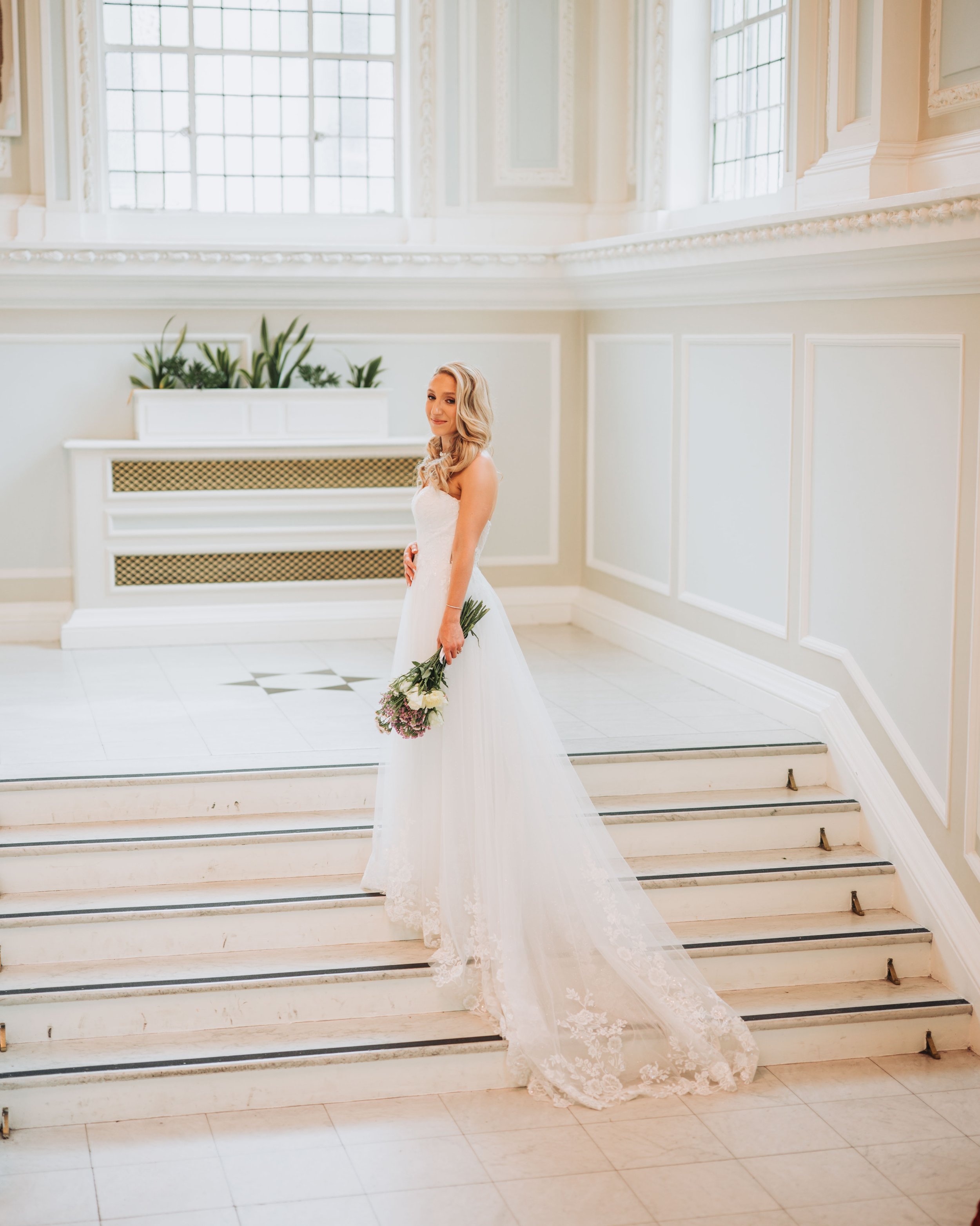 A bride holding a bouquet of flowers, standing on marble stairs in a bright, elegant hall with large windows and ornate architectural details.