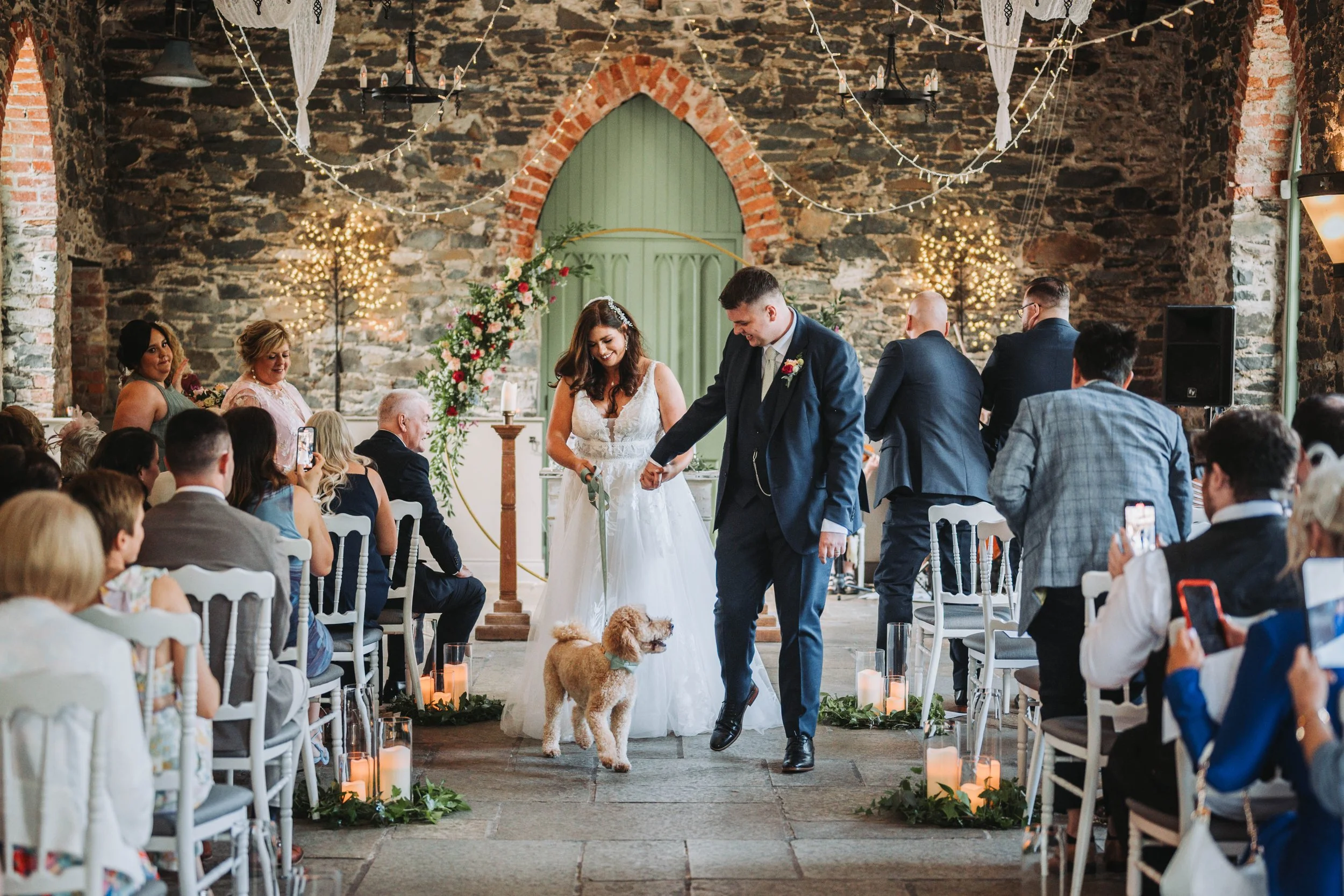 A couple walking a dog down the aisle during a wedding ceremony inside a rustic stone chapel decorated with candles, floral arrangements, and string lights.