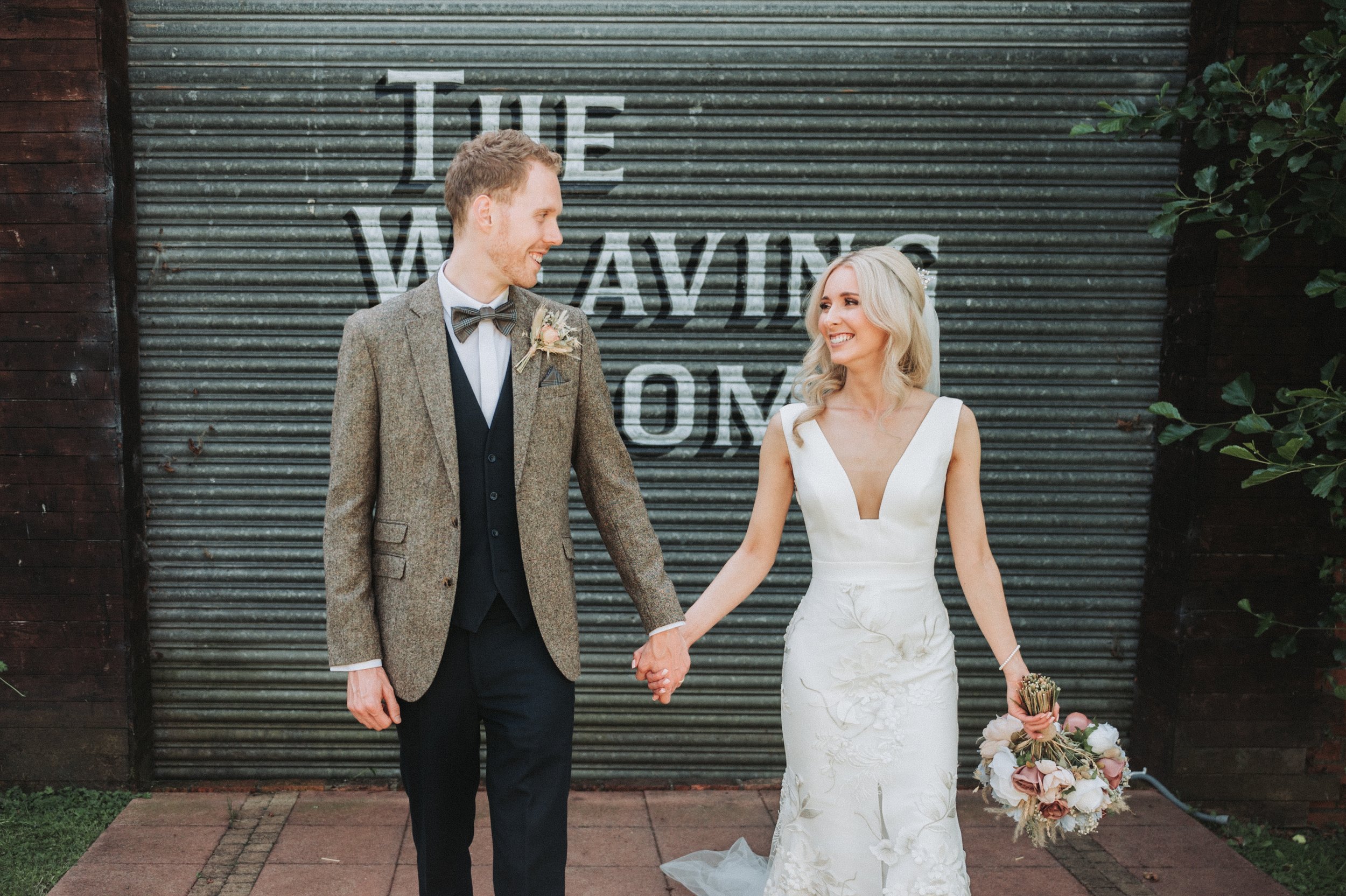 Bride and groom holding hands in front of "The Weaving Room" sign, bride in white dress with bouquet, groom in suit with bow tie.