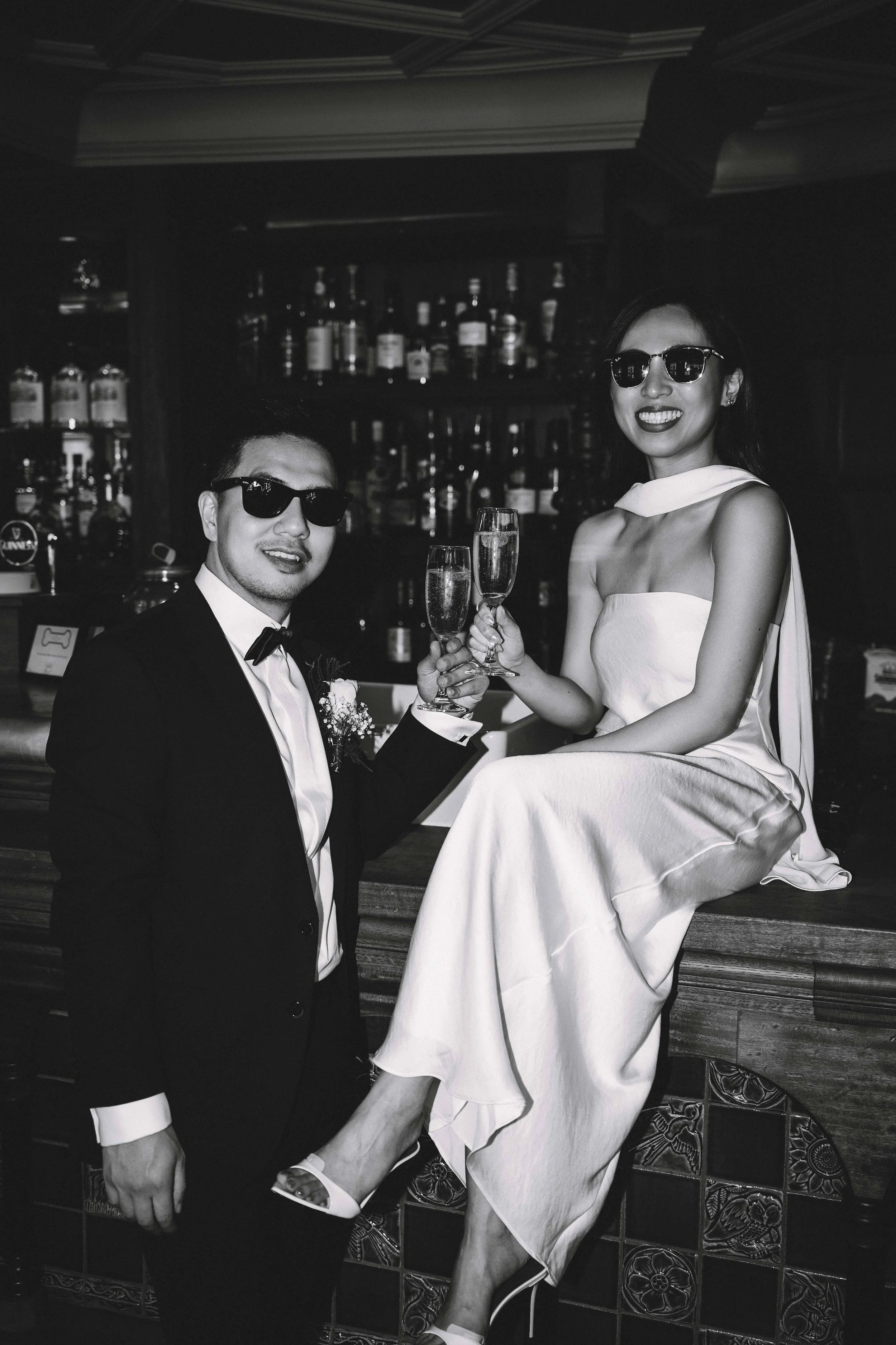 A black and white photo of a man and woman dressed in formal attire, wearing sunglasses, holding champagne glasses, celebrating at a bar.