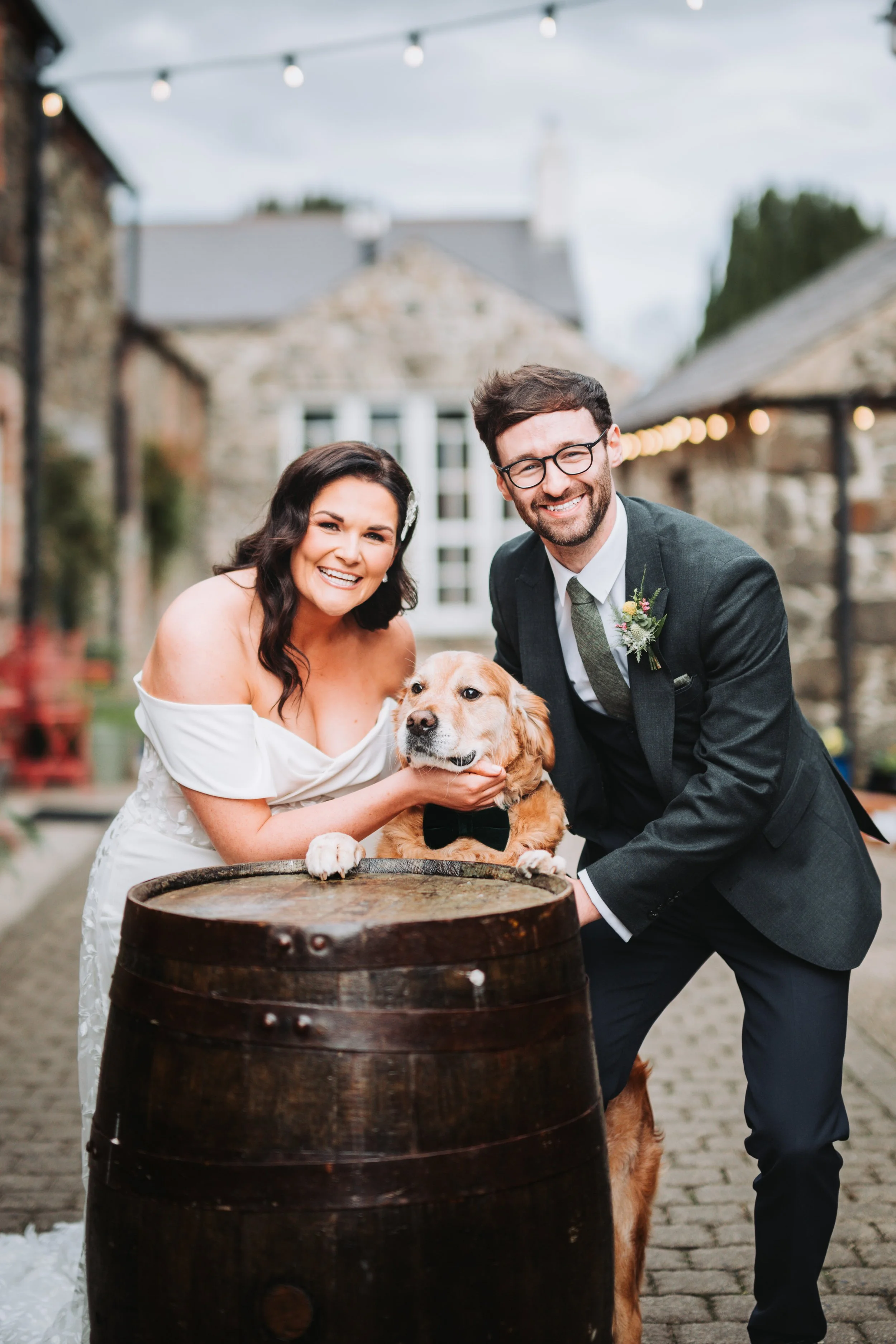 A wedding couple pose with their dog in an outdoor rustic setting, smiling at the camera.