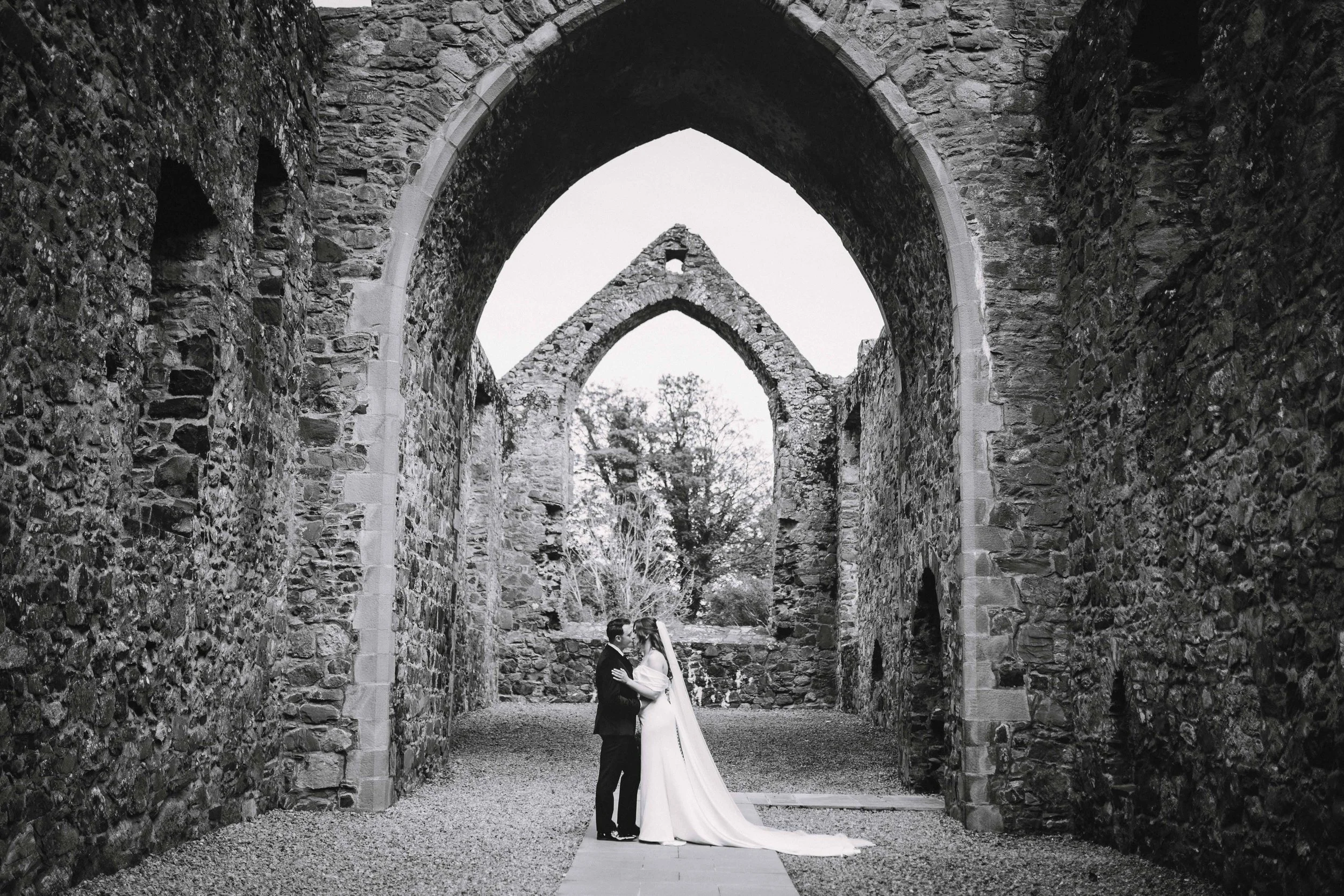 A black and white photo of a bride and groom standing close together, embracing under a large stone arch at an old stone ruin, with trees visible in the background.