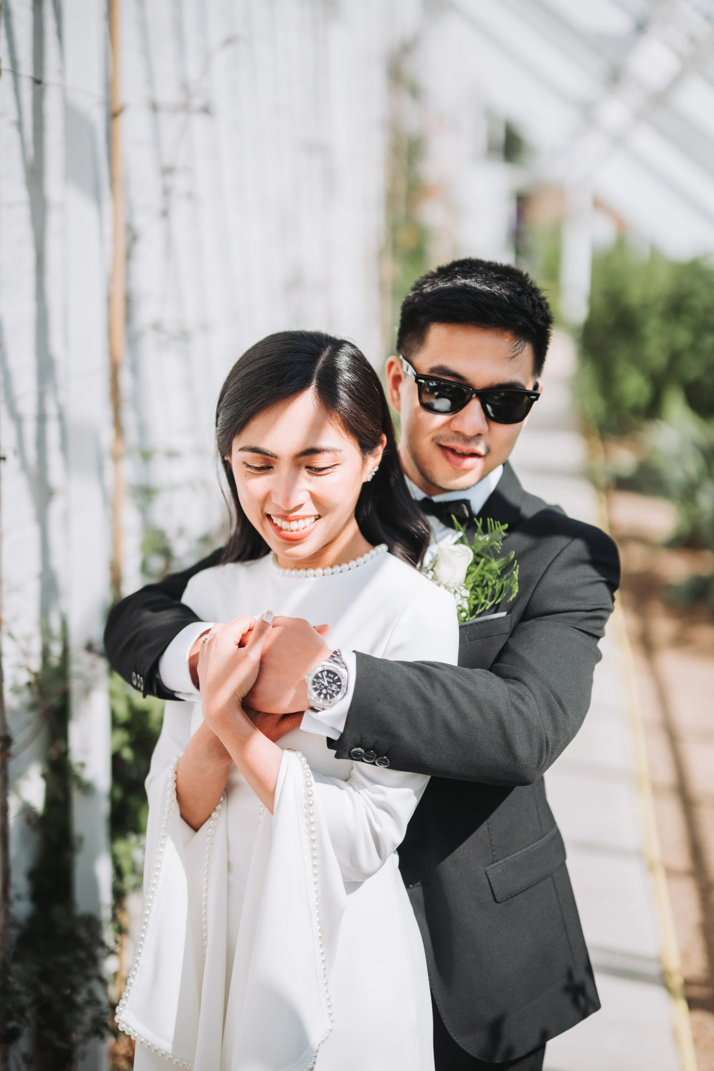 A couple in wedding attire sharing a joyful moment together inside a greenhouse or conservatory.