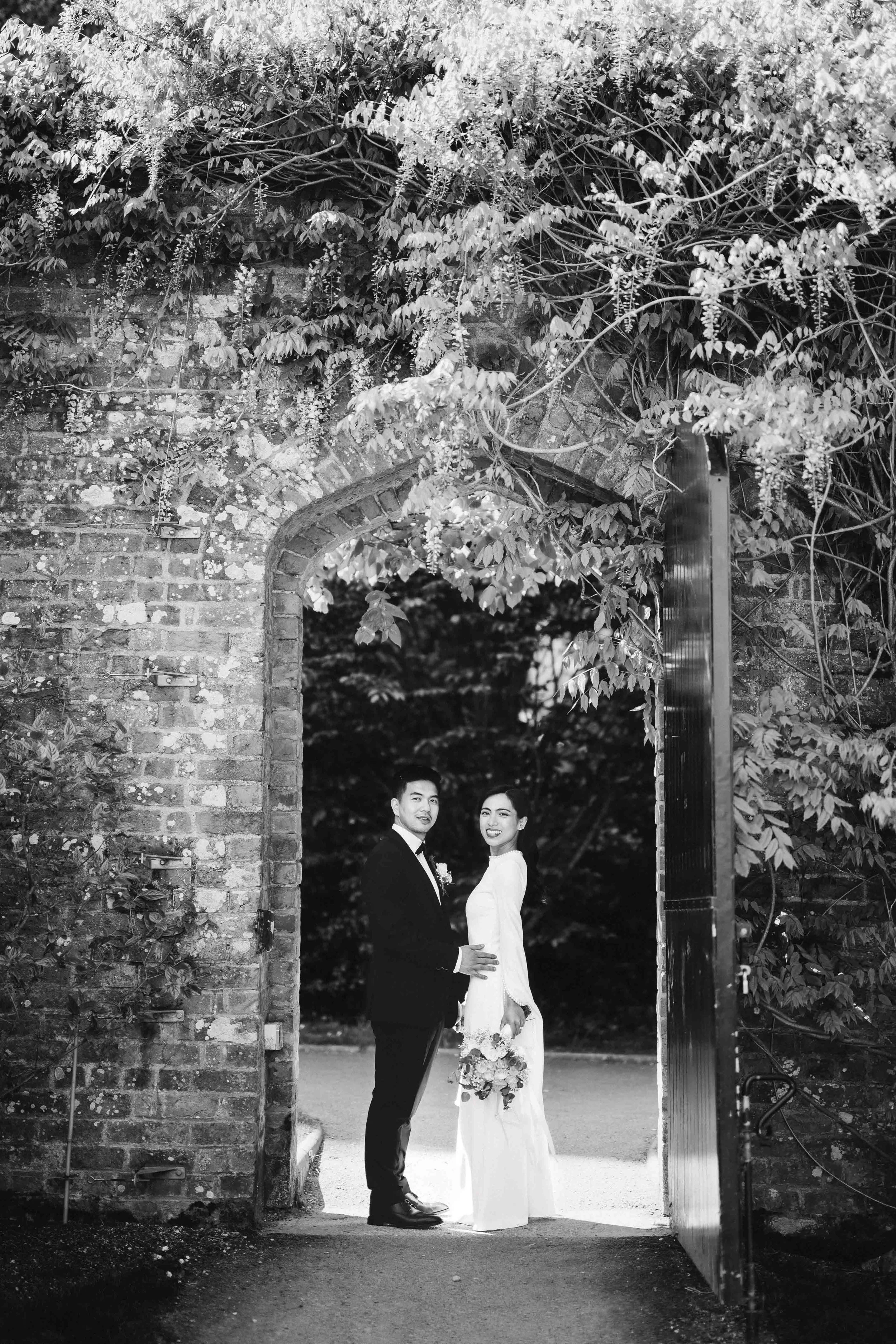 A black and white photo of a couple dressed in wedding attire, standing at an open gate framed by brick walls and overgrown foliage.
