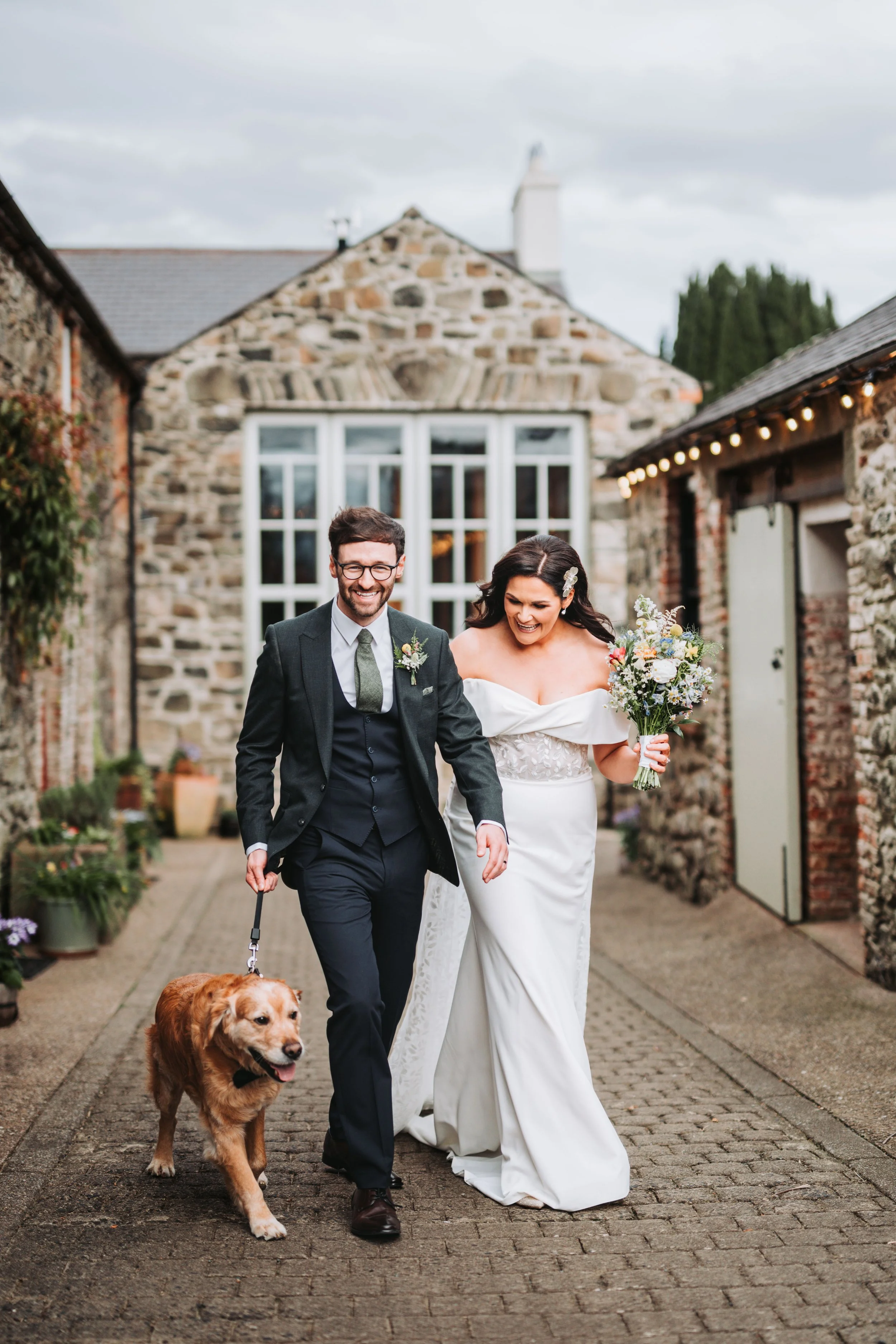 A newlywed couple walking outdoors with their dog, smiling and holding hands, in front of a rustic stone building with potted plants and string lights.