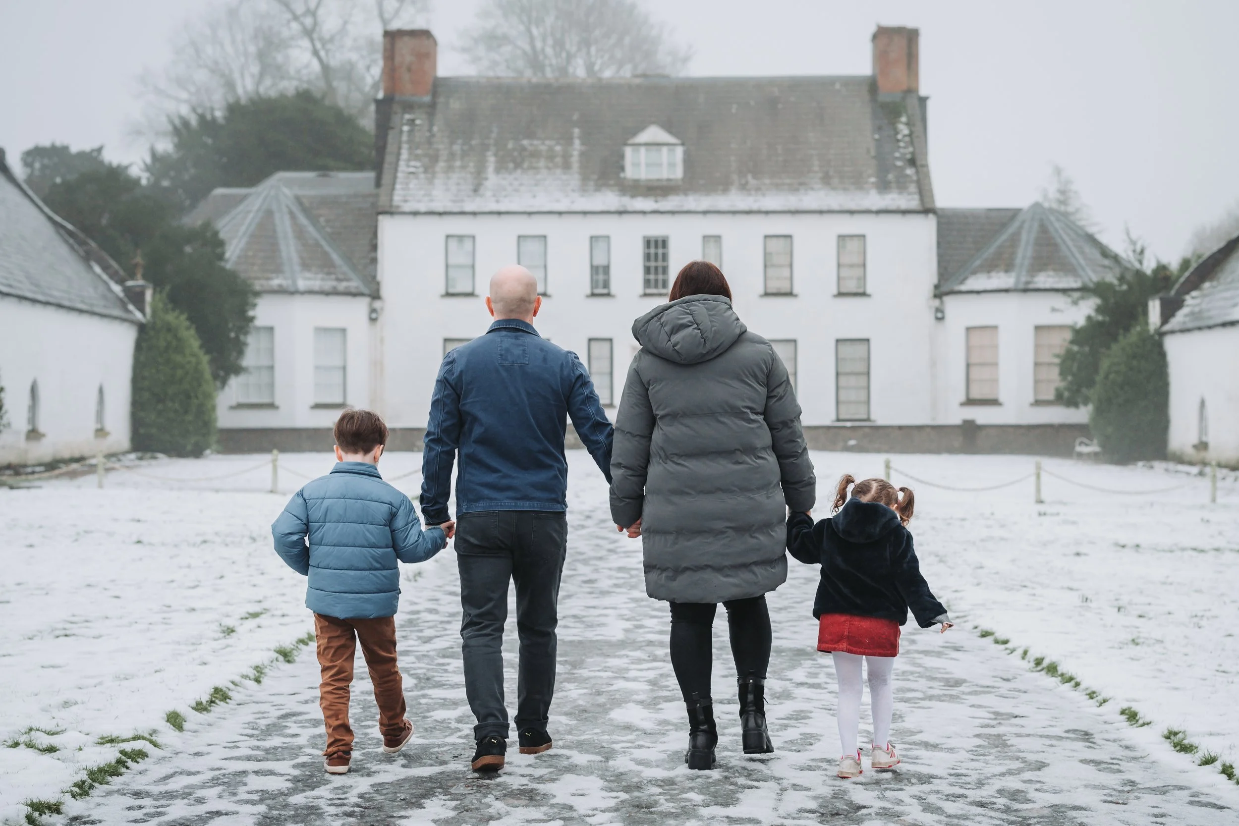 A family of four, including two children and two adults, walking hand in hand on a snow-dusted pathway toward a large white house in a winter landscape.