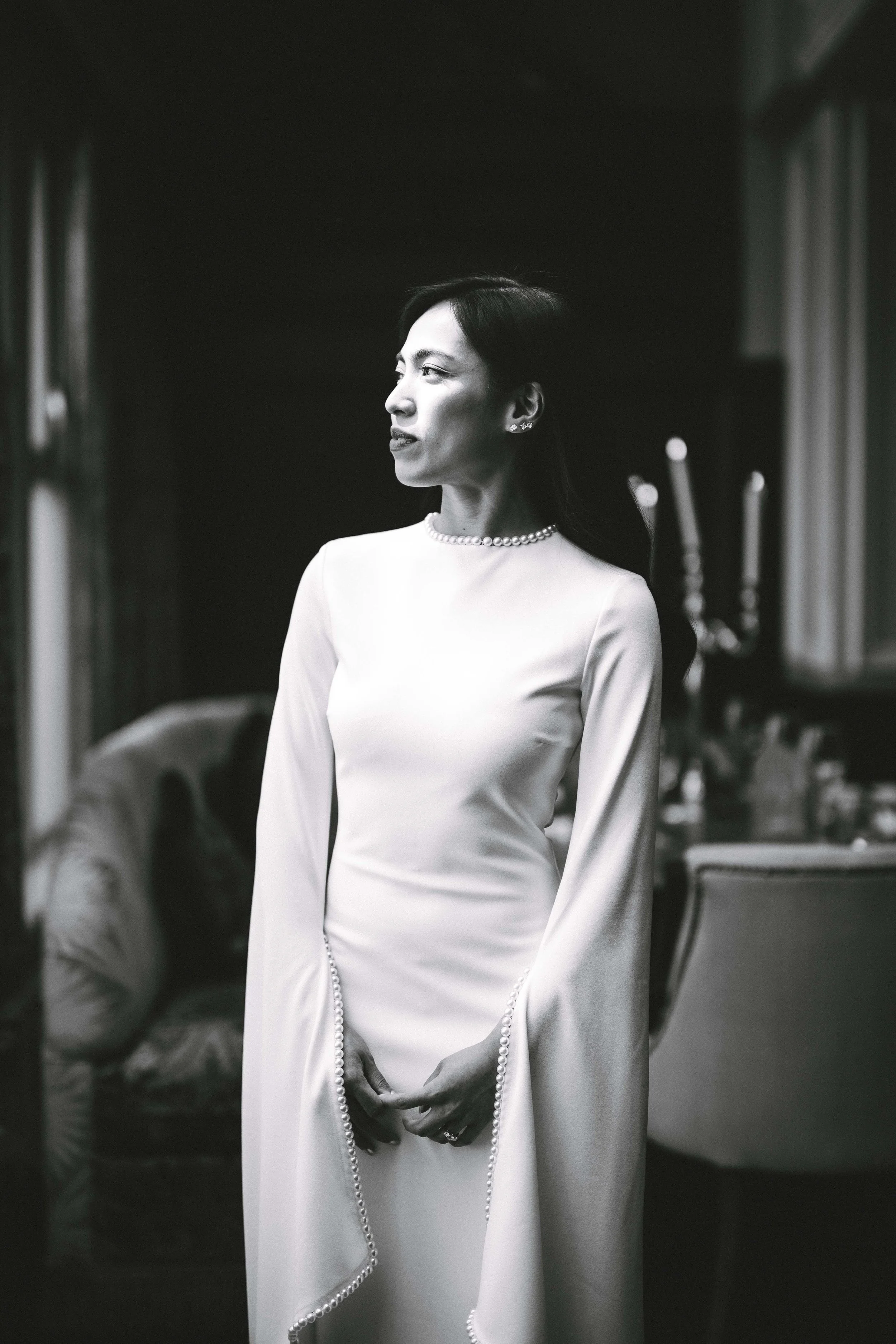 A woman in a long, elegant white dress with pearl accents, standing indoors with a contemplative expression, in a black and white photograph.