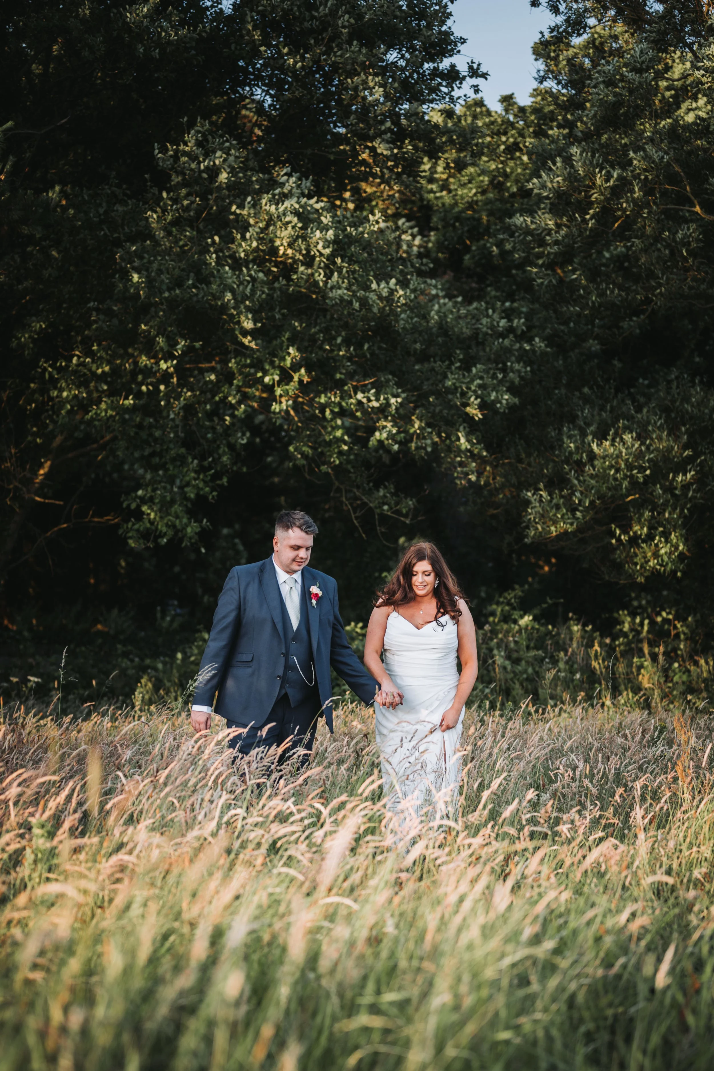 A newlywed couple holding hands and walking through a field of tall grass with trees in the background.