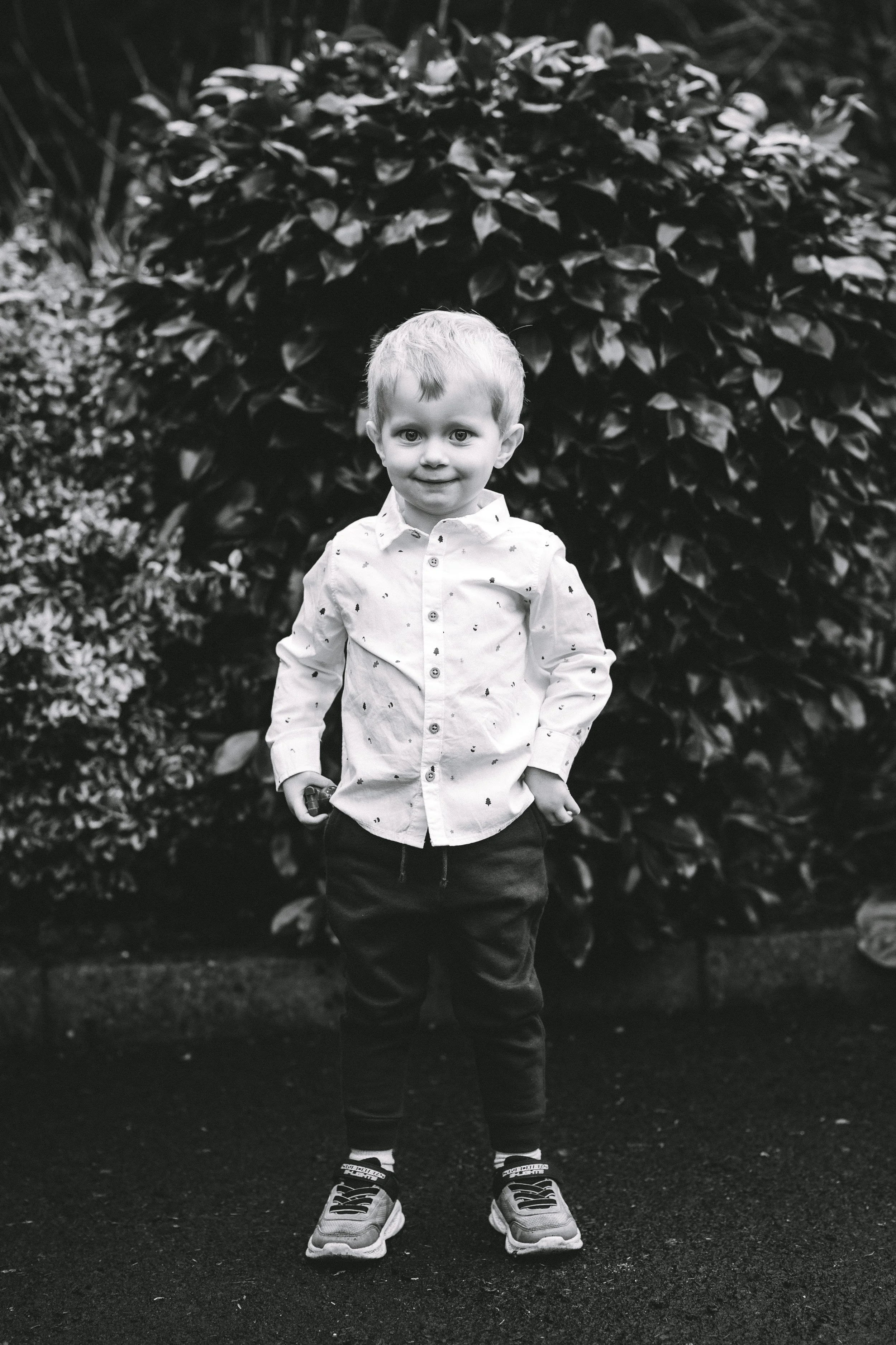A young boy with short hair standing outdoors in front of a bush, wearing a button-up shirt, dark pants, and sneakers.