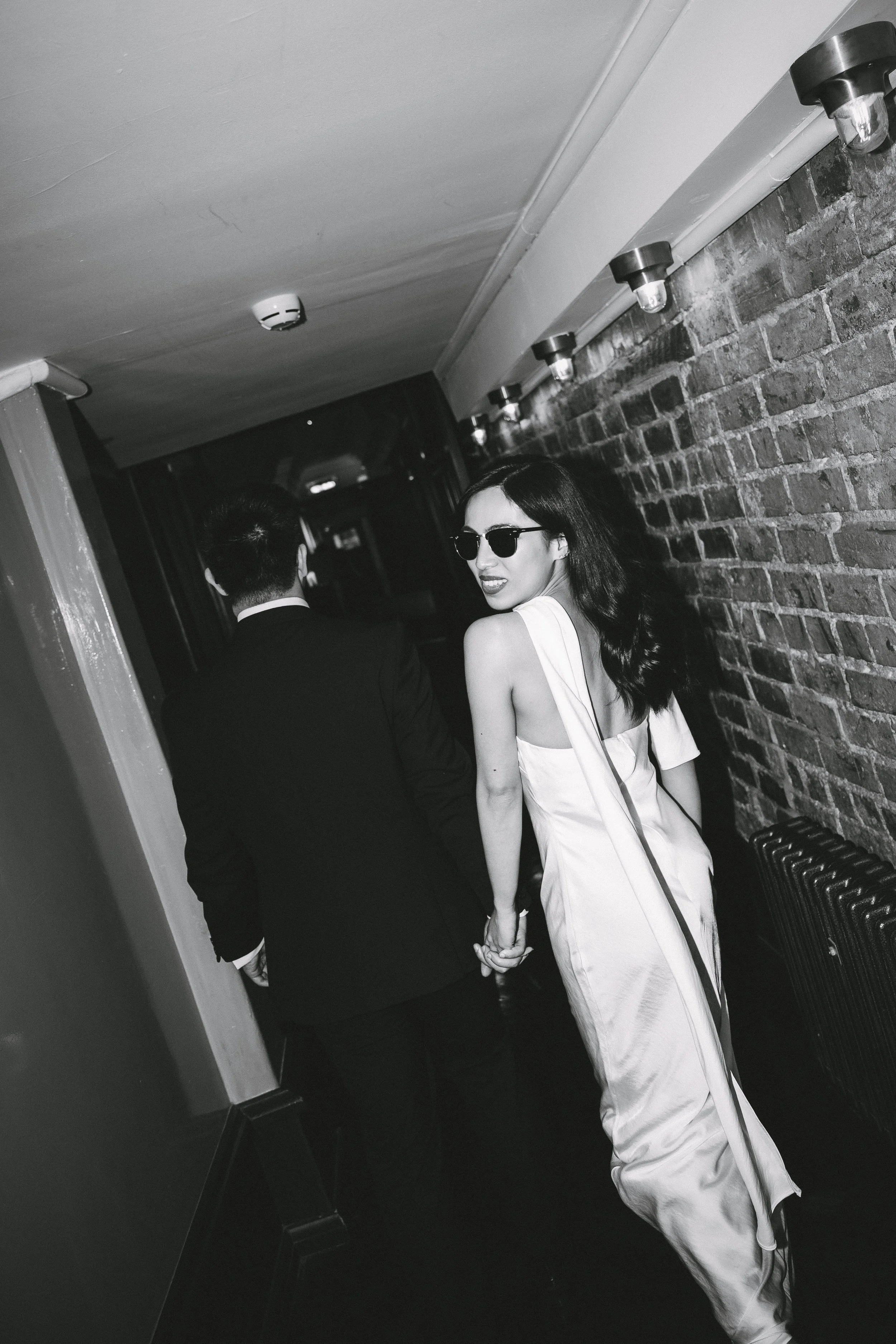 A black and white photo of a woman in a satin dress and sunglasses holding hands with a man in a suit, walking down a dimly lit corridor with a brick wall on one side.