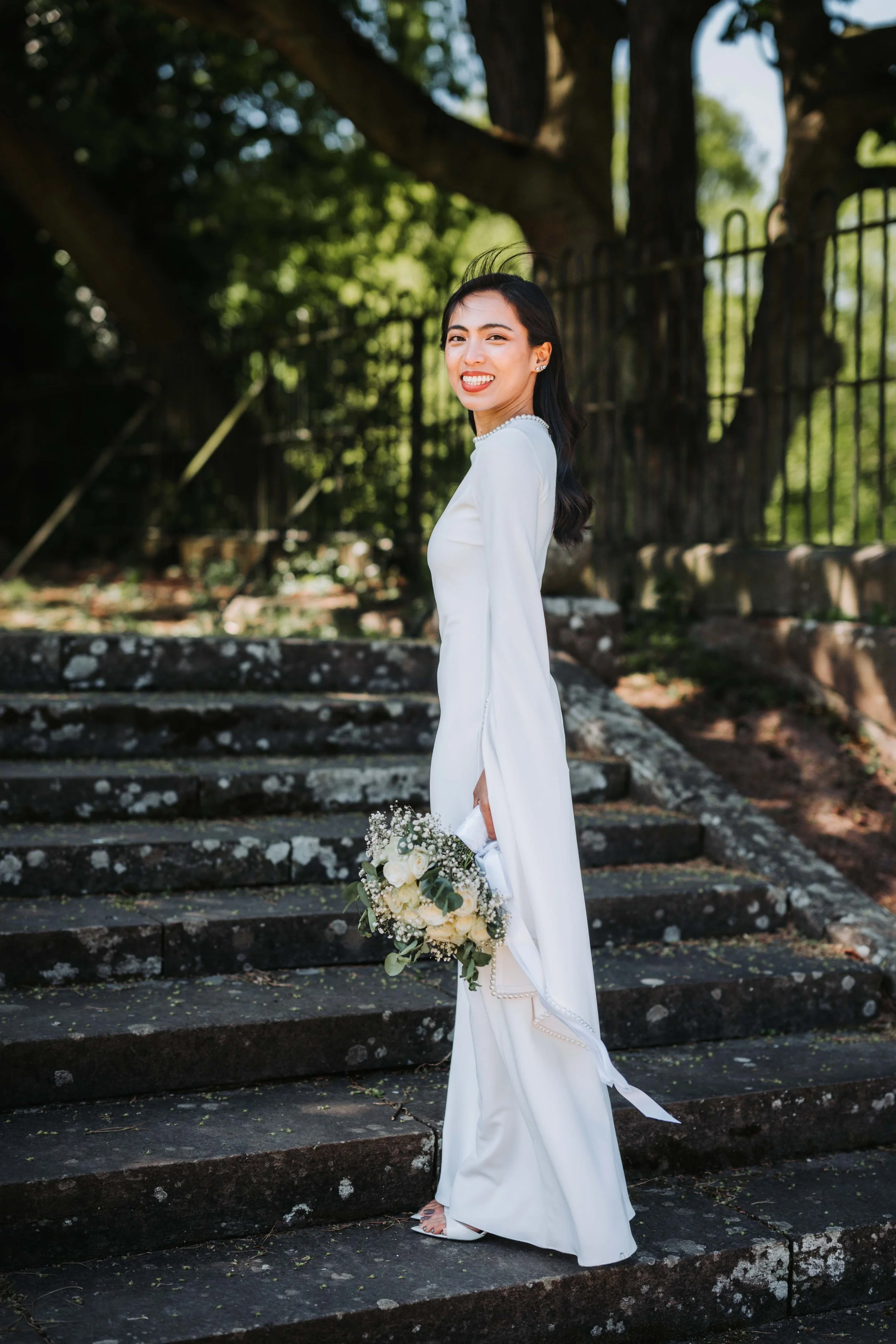 A woman in a white dress holding a bouquet of white roses and greenery, standing outdoors on stone steps with a large tree and metal fence in the background.