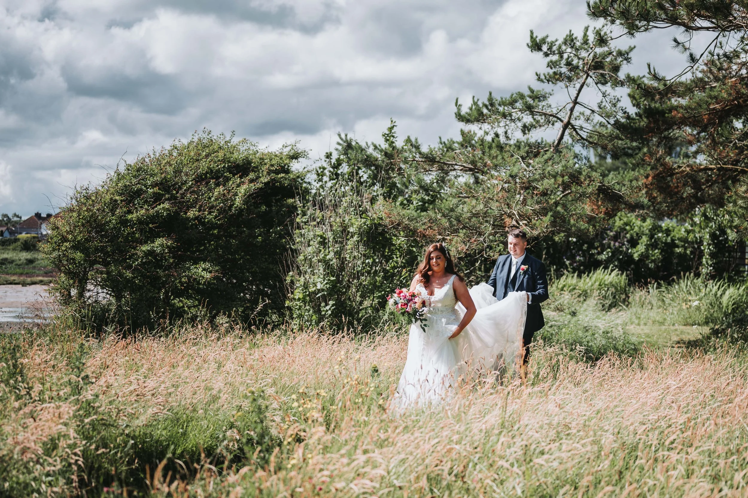 Bride and groom walking through a grassy field during daytime. The bride is holding a bouquet of pink and white flowers, wearing a white wedding dress. The groom is in a dark suit, holding the bride's dress. There are green trees and bushes in the ba