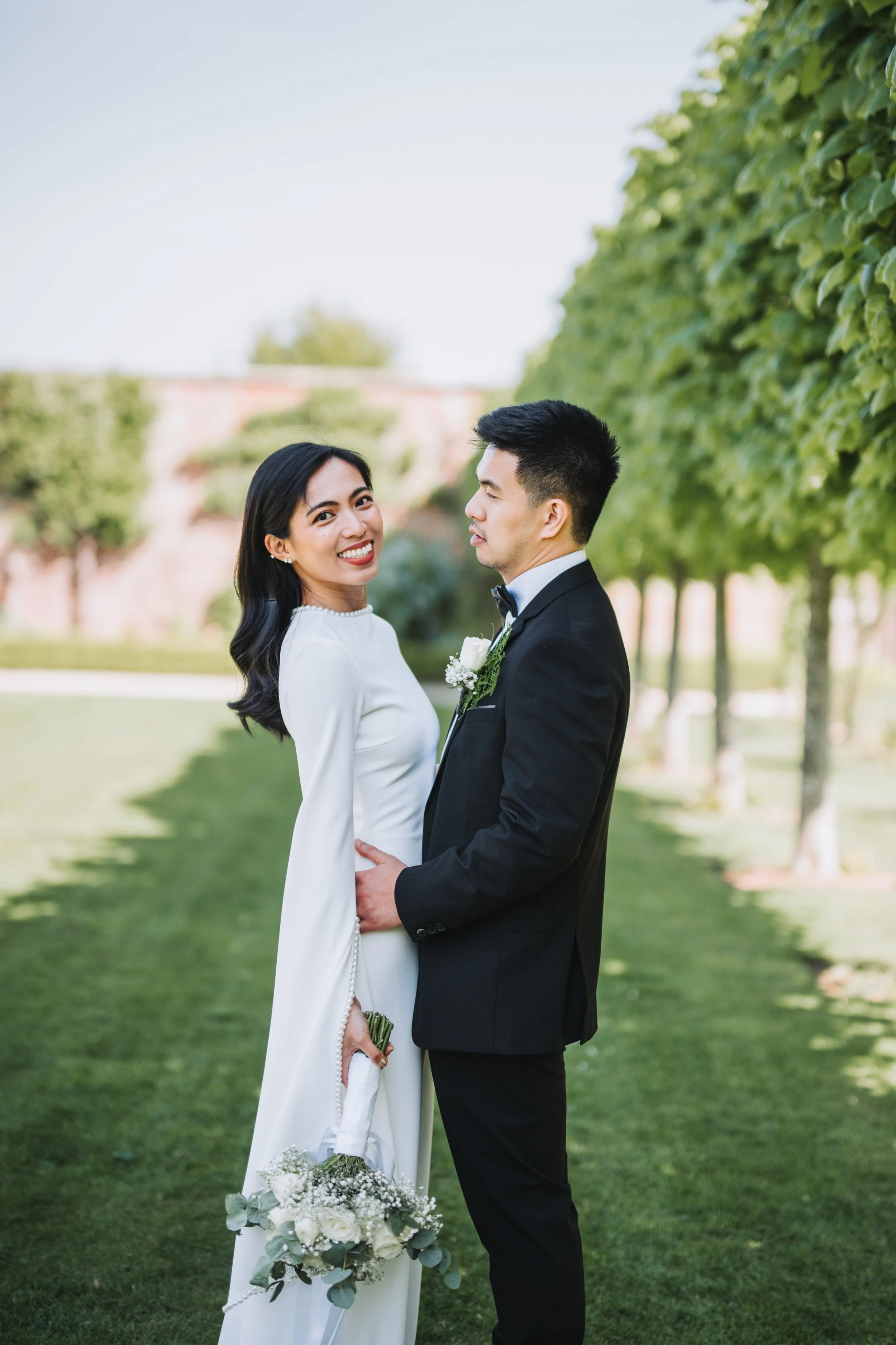 A newlywed couple outdoors on their wedding day, the woman holds a bouquet and smiles at the camera while the man, dressed in a tuxedo, gently holds her around the waist amidst green trees.