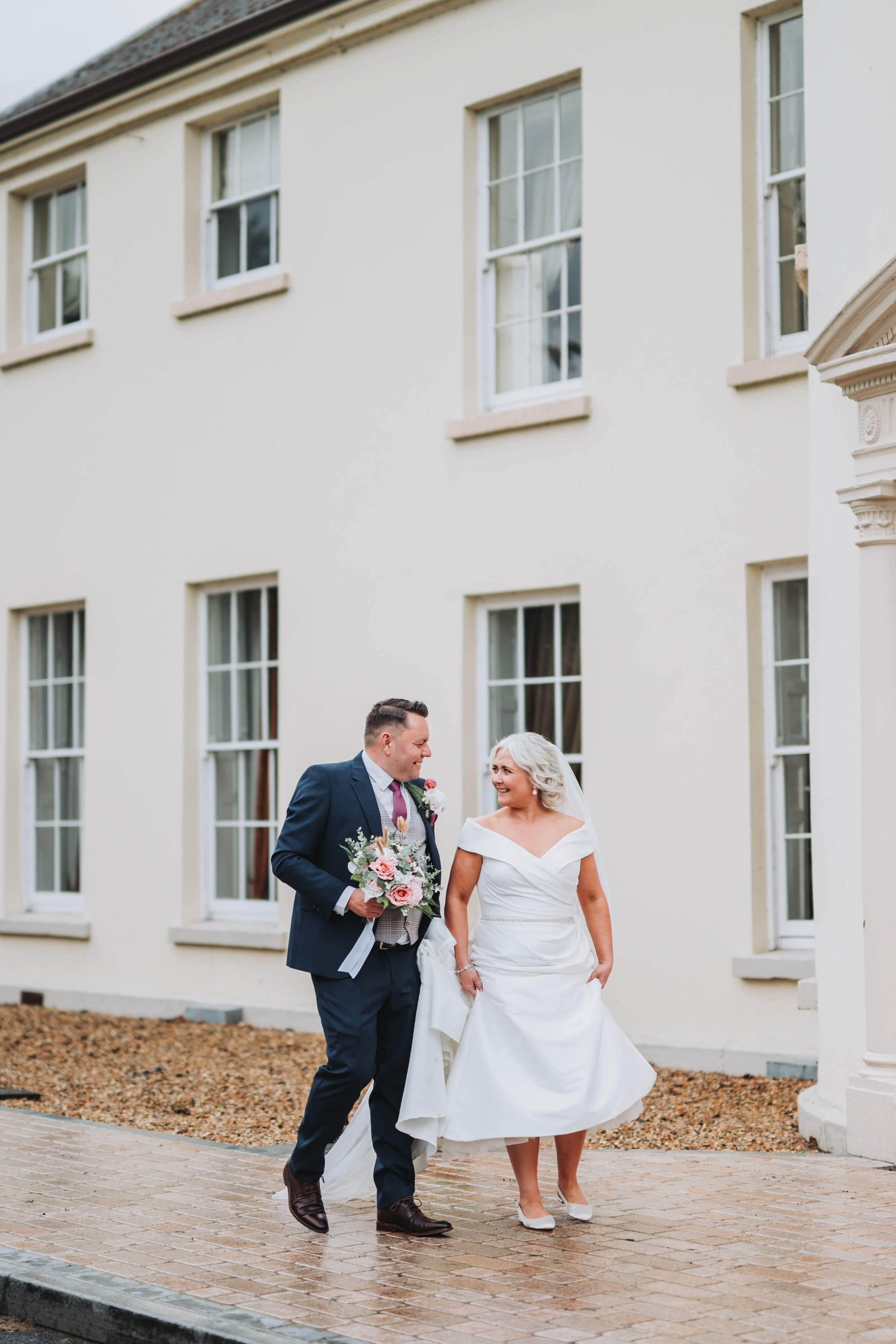 A couple dressed in wedding attire walking outdoors in front of a white building, smiling at each other. The groom holds a bouquet of flowers.